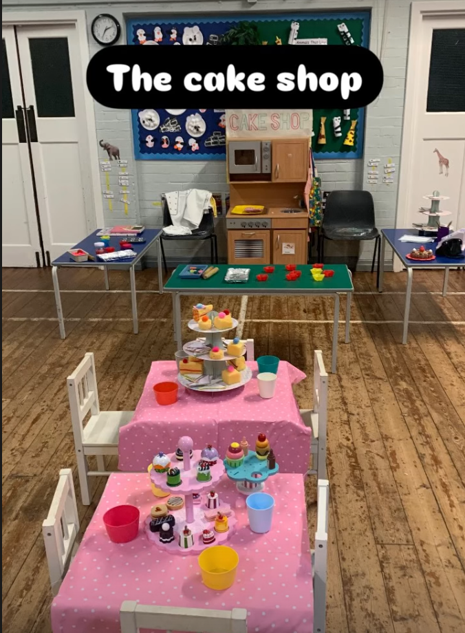 A classroom setup with a cake shop pretend play area. There are two small tables with pink polka dot tablecloths, decorated with toy cupcakes, cakes, and pretend desserts. In the background, there's a play kitchen, and on the walls are children’s decorative artwork and a bulletin board.