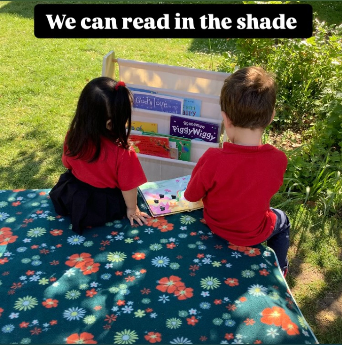 Two children sitting on a floral blanket outdoors, reading books from a portable bookshelf, with a sign above that says 'We can read in the shade'.