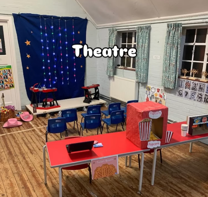A children's theater setup with a blue starry backdrop and dangling string lights, stage with toy scooters, and small blue chairs facing the stage. There's a red table with a popcorn machine, cups, and a sign that says 'Ticket Booth'. The room has windows with green floral curtains and wooden walls.