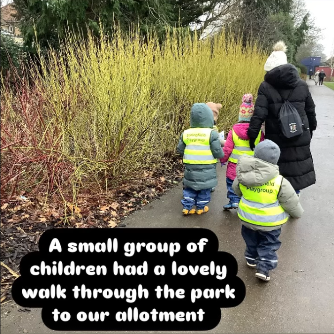 A small group of children and an adult walking through a park on a paved path, surrounded by tall bushes and trees, with children wearing yellow reflective vests. The children are dressed warmly with jackets, hats, and walking shoes.