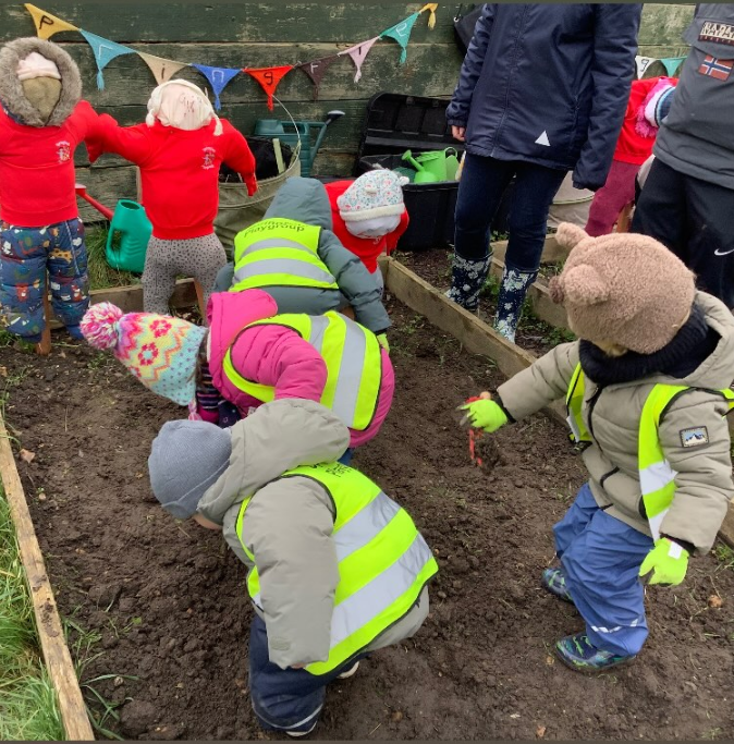 Children in warm clothes and high-visibility vests planting or harvesting crops in a garden bed.