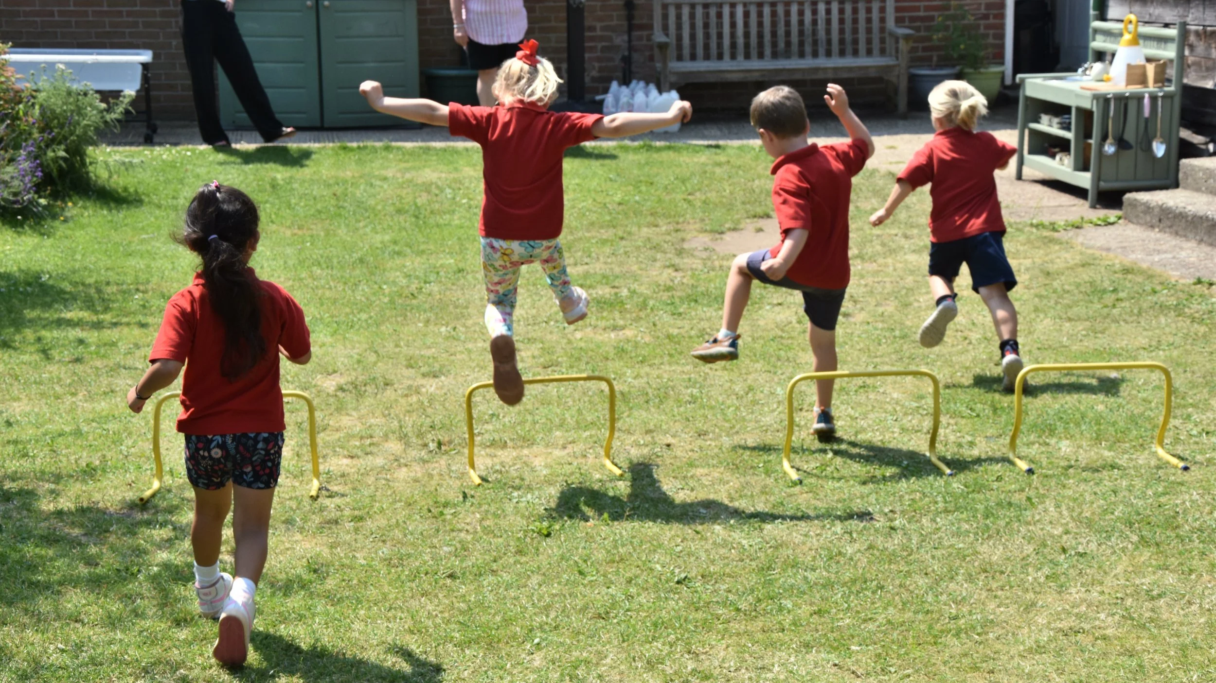 Four children in red shirts playing outdoors on grass, jumping over yellow hurdles, with a girl in a patterned skirt observing, and adults in the background near a garden and patio area.