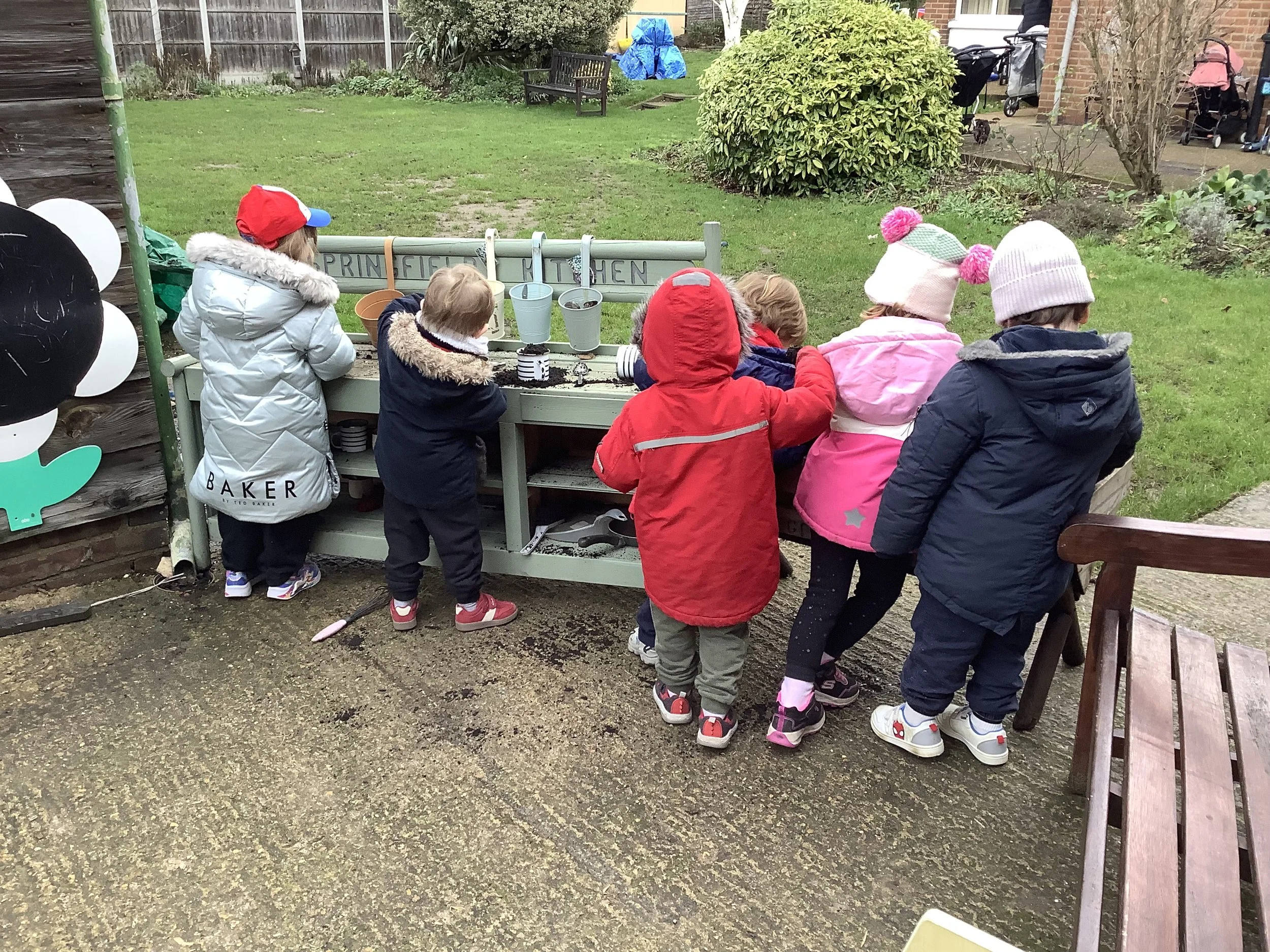 Seven children dressed in coats and hats are playing at a designated outdoor play area, which includes a green wooden kitchen set with pots, pans, and utensils, on a damp concrete surface in a garden setting with grass, bushes, and houses in the background.