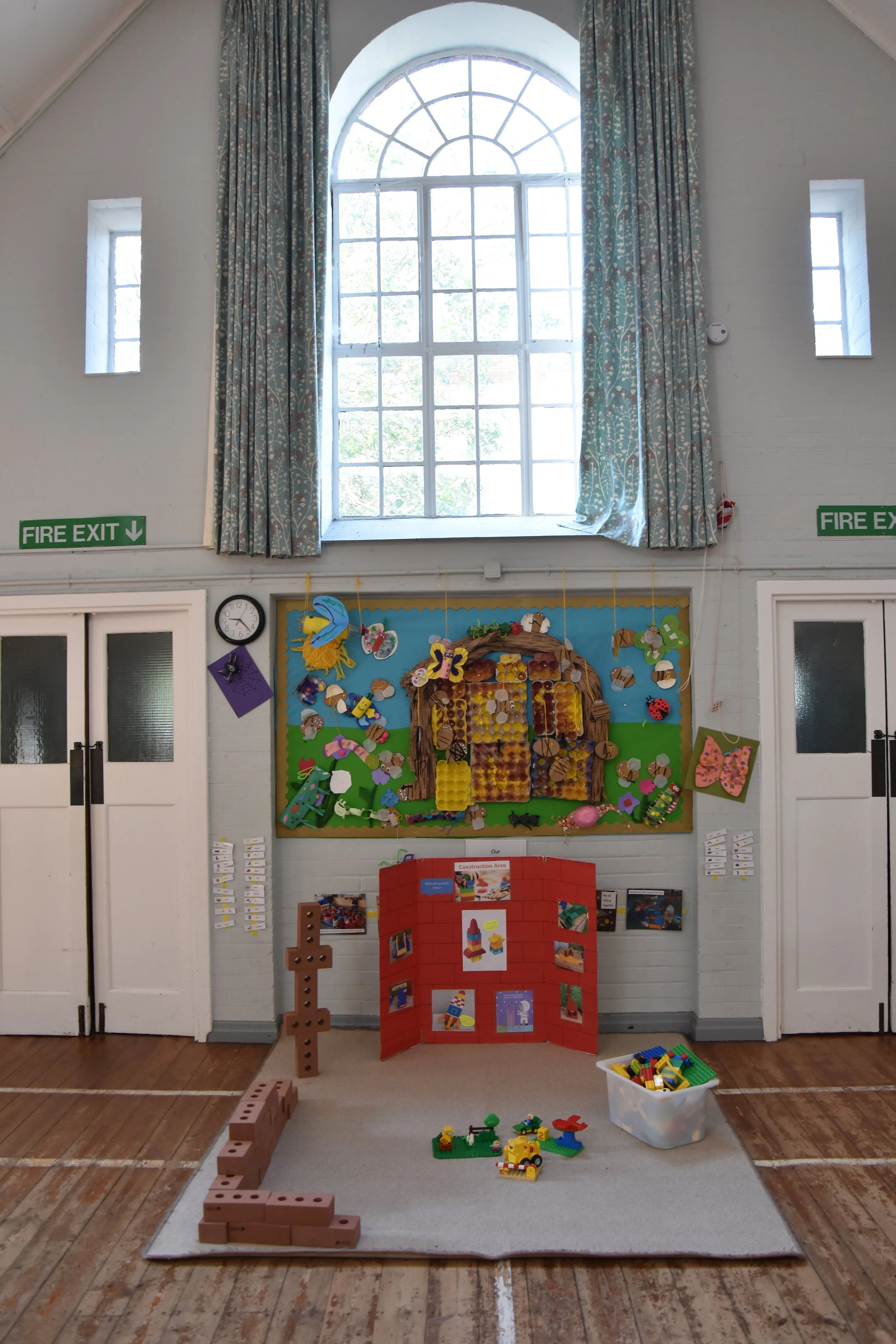 Classroom display area with a construction theme, featuring building blocks arranged to form a tower, a display board with animal and nature crafts, and a container of LEGO bricks on the floor. There are two white doors with frosted glass and two fire exit signs, with a large arched window and smaller windows letting in natural light.