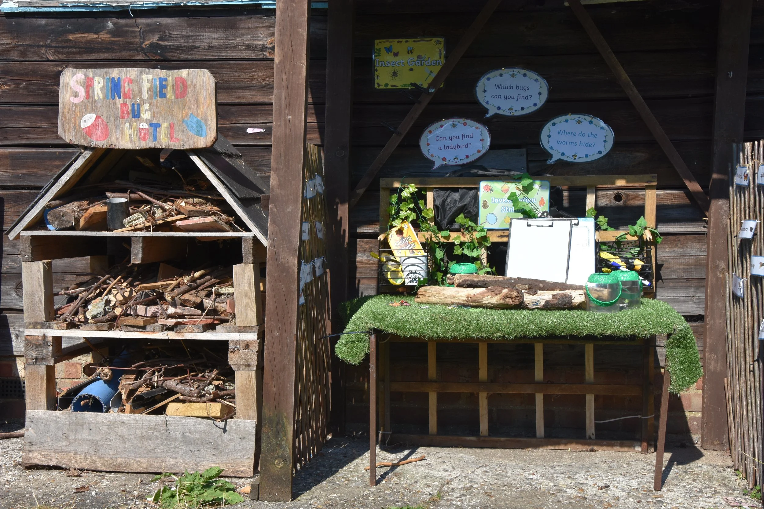 Outdoor educational station with a sign reading 'Springfield Bus & Hotel' on a wooden structure. To the right, a display with signs about bugs, ladybirds, and worms, along with a clipboard, containers, and a small log on a table covered with artificial grass. Behind the display, there is a wooden wall with decorative signs.