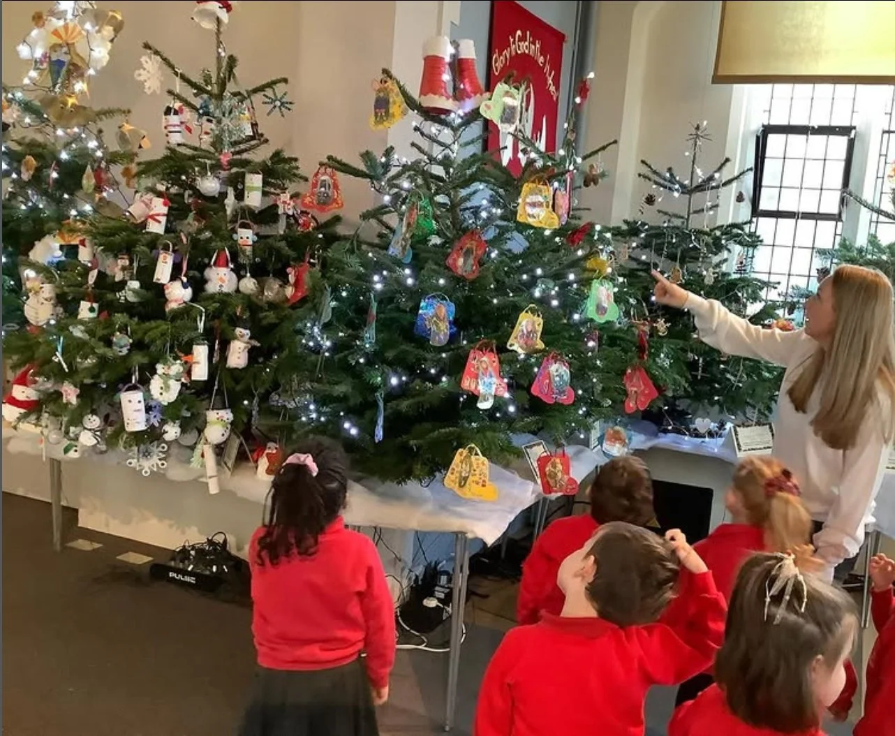 A teacher and a group of children in red uniforms school uniforms are gathered around a decorated Christmas tree with ornaments, lights, and drawings. The teacher is pointing to the tree, showing it to the children inside a classroom or community space.