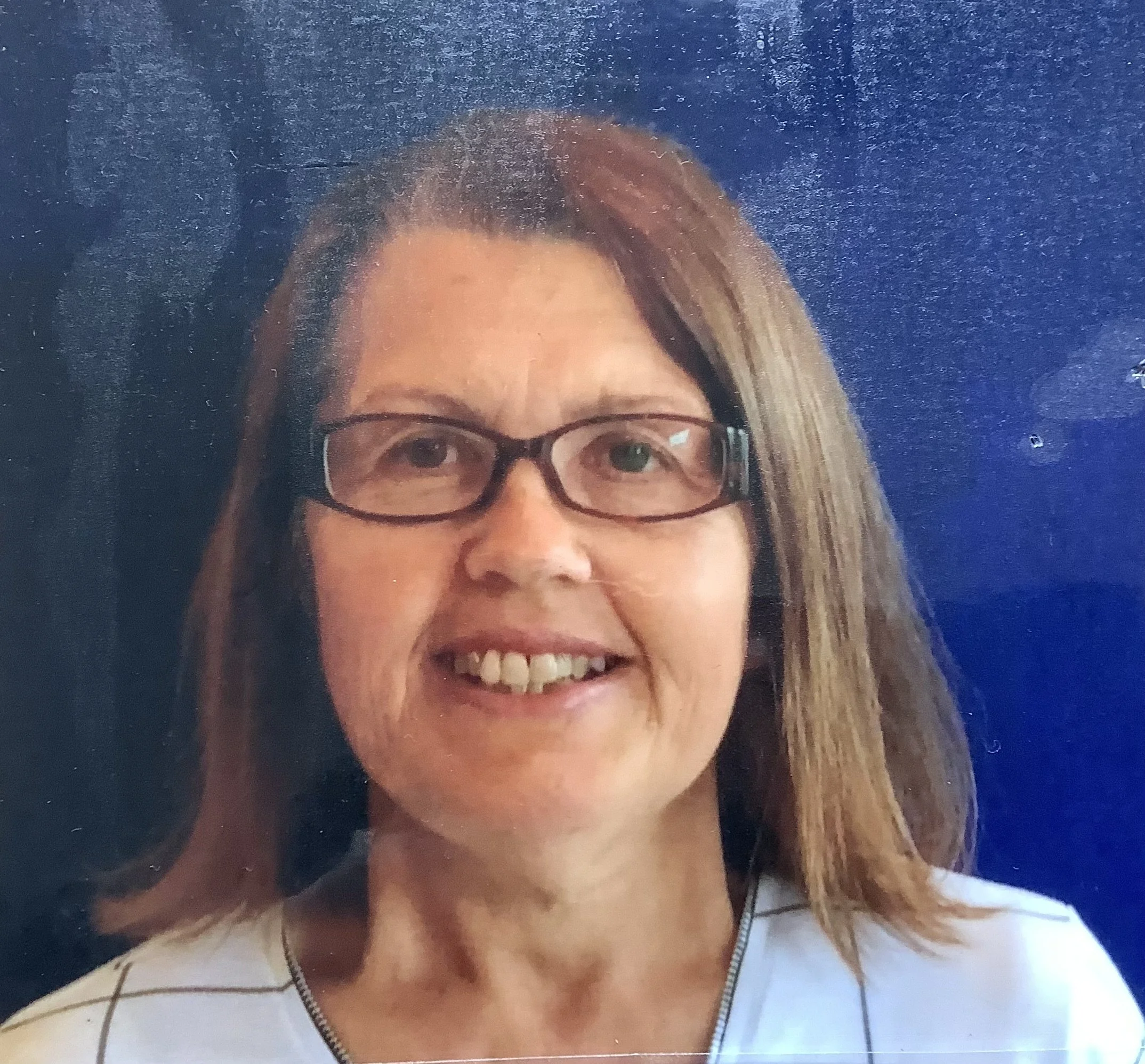 Close-up of a smiling woman with glasses, long hair, and a white shirt, standing in front of a dark blue background.