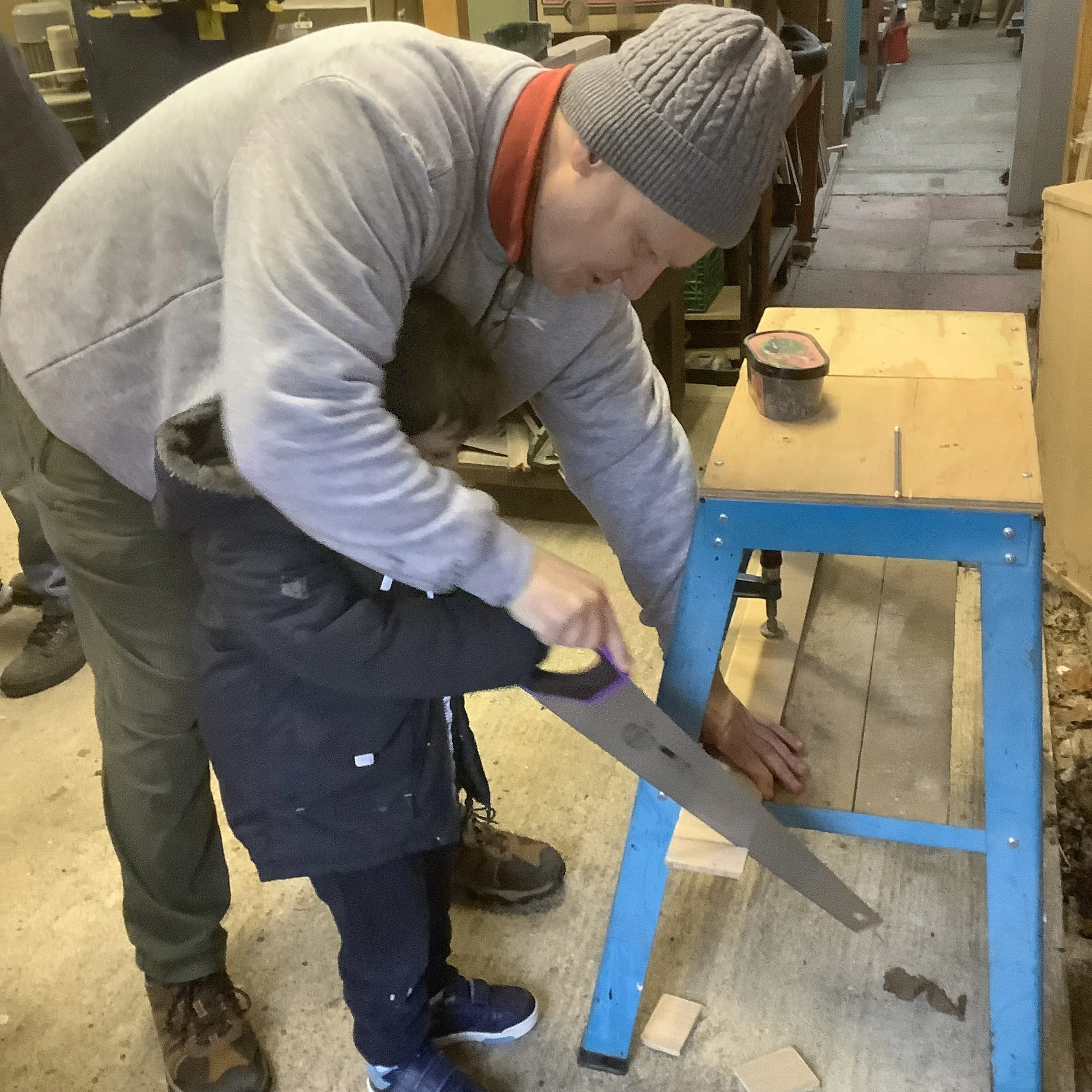 An adult male helping a child with a handsaw in a woodworking shop. The adult is guiding the child's hand as they cut a piece of wood on a workbench. Both are wearing warm clothing, and there are tools and supplies in the background.