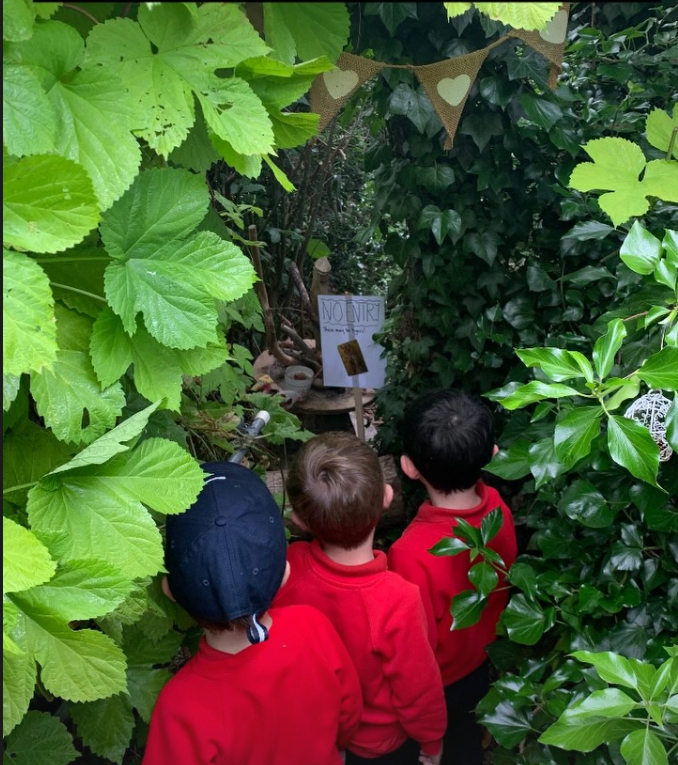 Three children in red shirts are observing a small, decorative outdoor shrine adorned with green foliage, a 'No NUK' sign, and small objects or offerings, set amidst dense greenery.