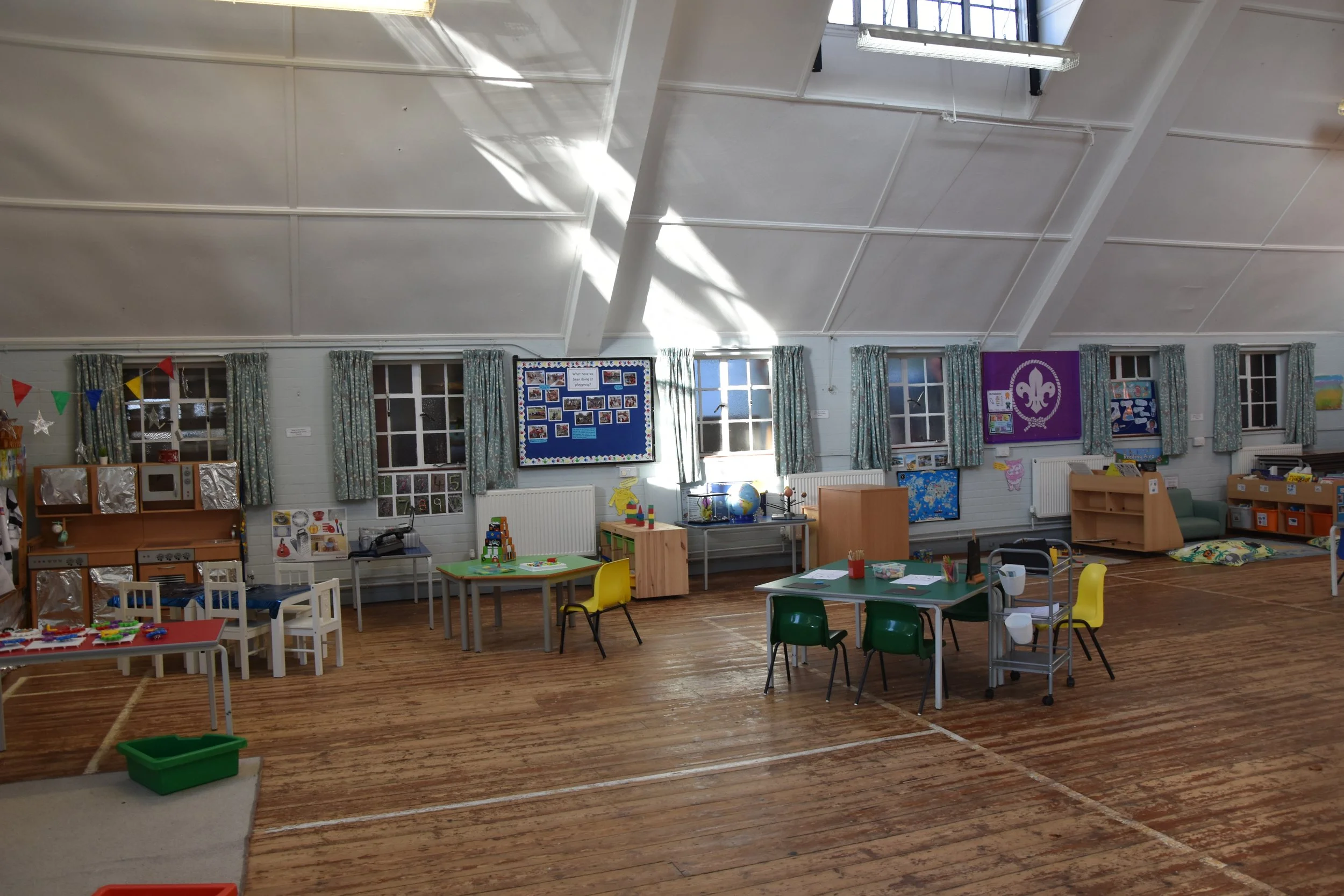 A classroom with wooden flooring, several small tables and chairs, decorated walls, and windows with curtains. There are various educational materials and decorations on the walls, including a bulletin board, posters, and a flag. The room is well-lit, with sunlight streaming through the windows.