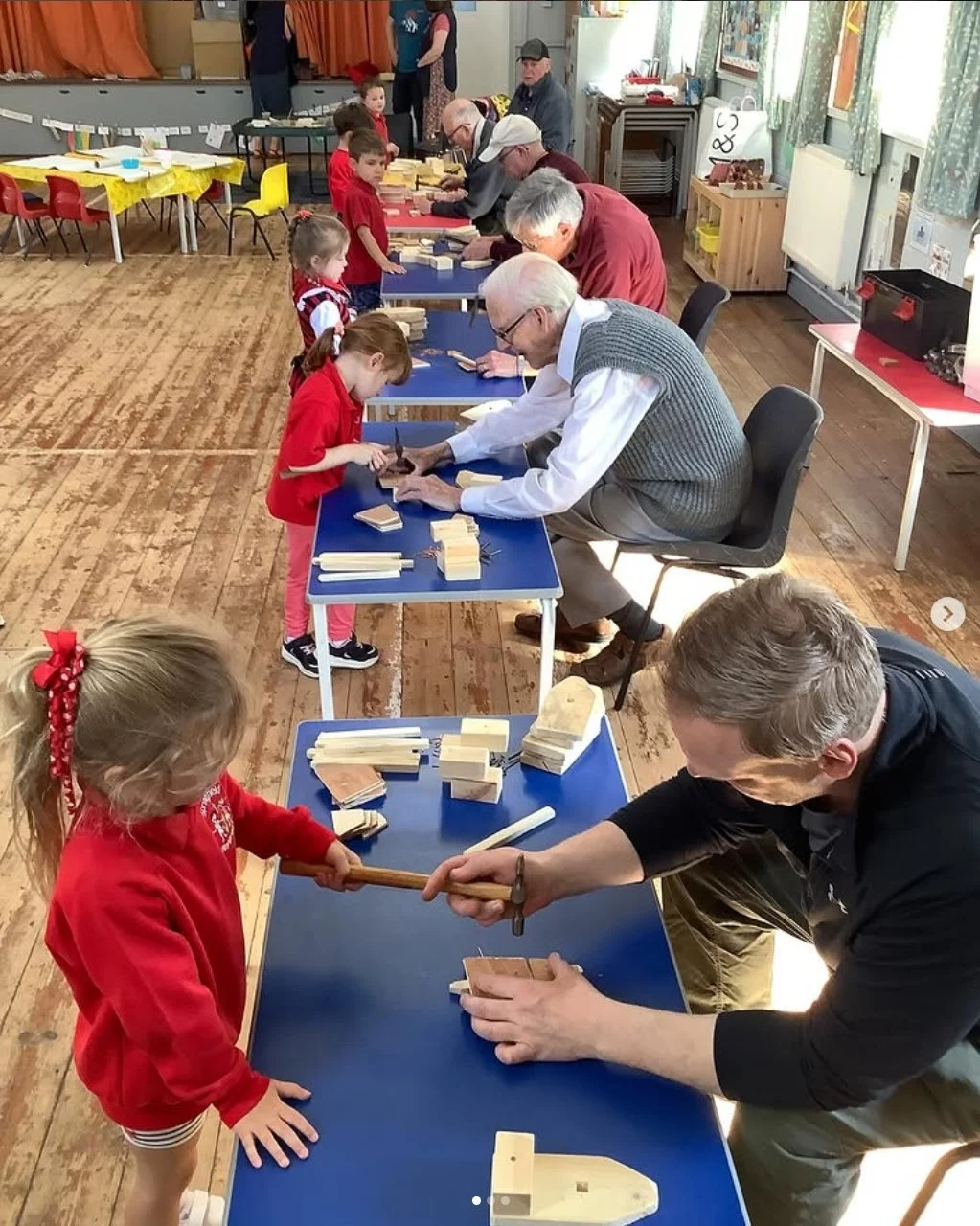 Adults and children playing with wooden blocks at tables in a community room, engaging in woodworking activities.