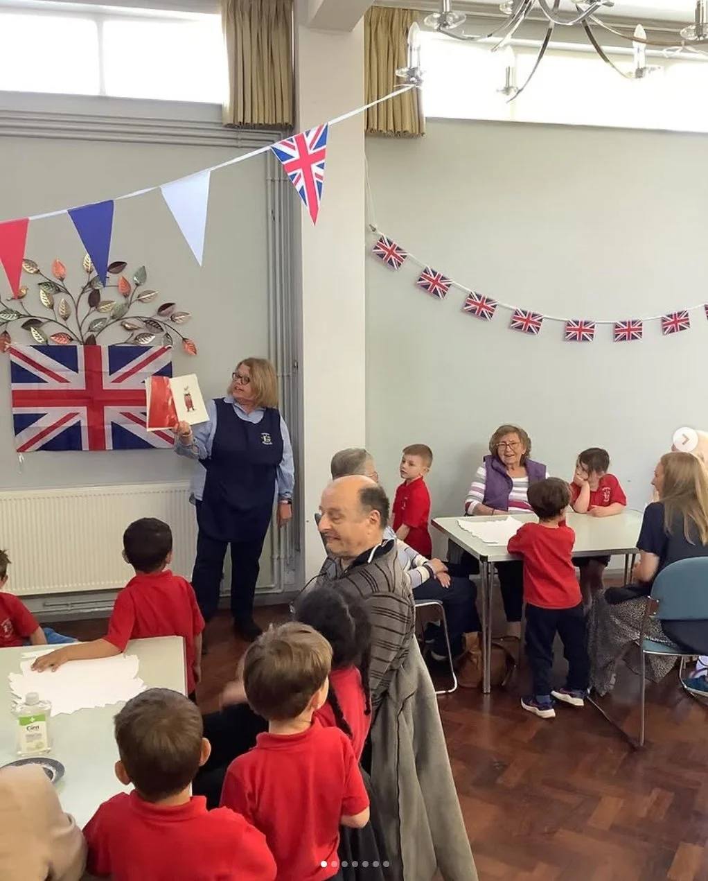 A group of children and adults in a room decorated with British flags, one adult woman holding a book while standing near a wall with a large Union Jack and a tree art display, others sitting or standing at tables, some children wearing red shirts, celebrating a UK-themed event.