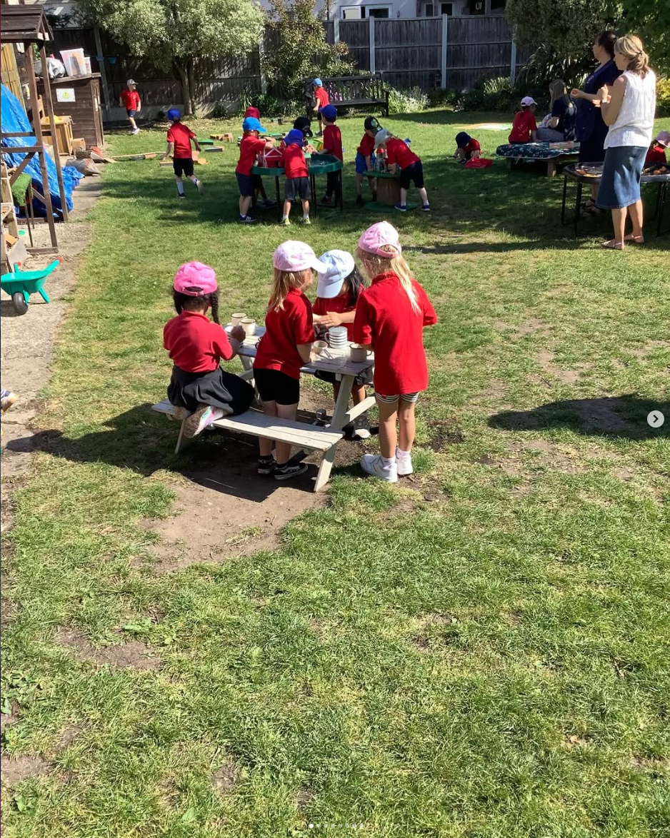 Children in red uniforms and hats playing outdoors on a grassy area, with some sitting at a small picnic table and others engaging in activities near a fence and a shed.