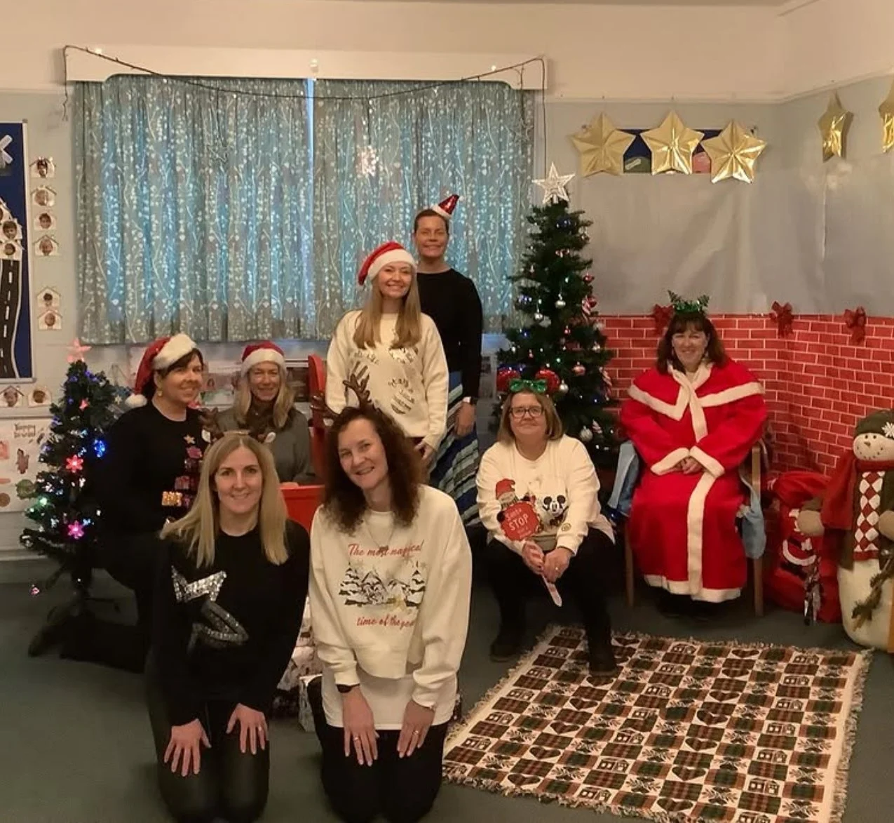 Group of nine women celebrating Christmas indoors, some wearing Santa hats, with decorated Christmas trees and festive decorations around, including gold stars and a snowman. They are smiling, sitting and standing in front of a window with blue curtains, a Christmas tree, and holiday garlands.