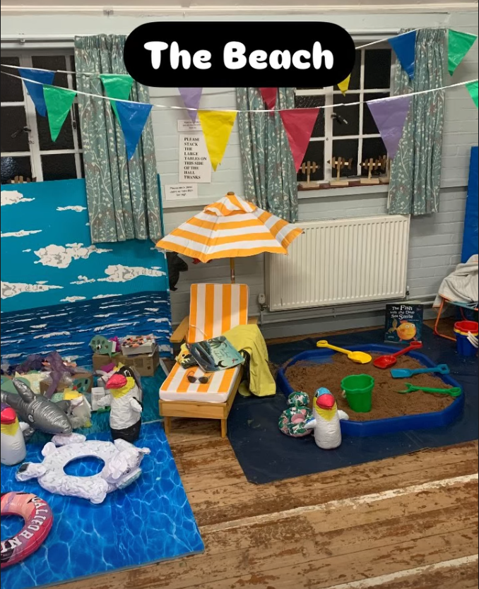 A beach-themed play area with a painted backdrop of ocean waves and clouds, colorful bunting flags, a striped beach chair and umbrella, a small sandbox with toy shovels and buckets, inflatable pool floats, and stuffed animals resembling penguins, creating a mini beach scene.
