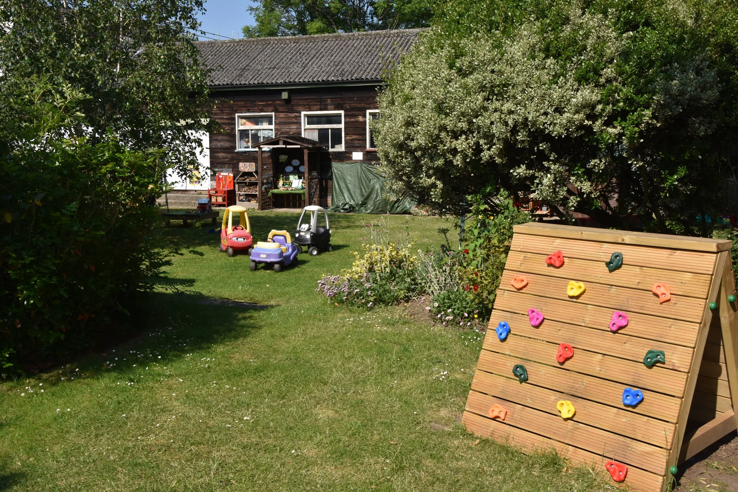 A backyard garden with a wooden house, colorful toy cars, a rock climbing wall, and lush green trees and plants.