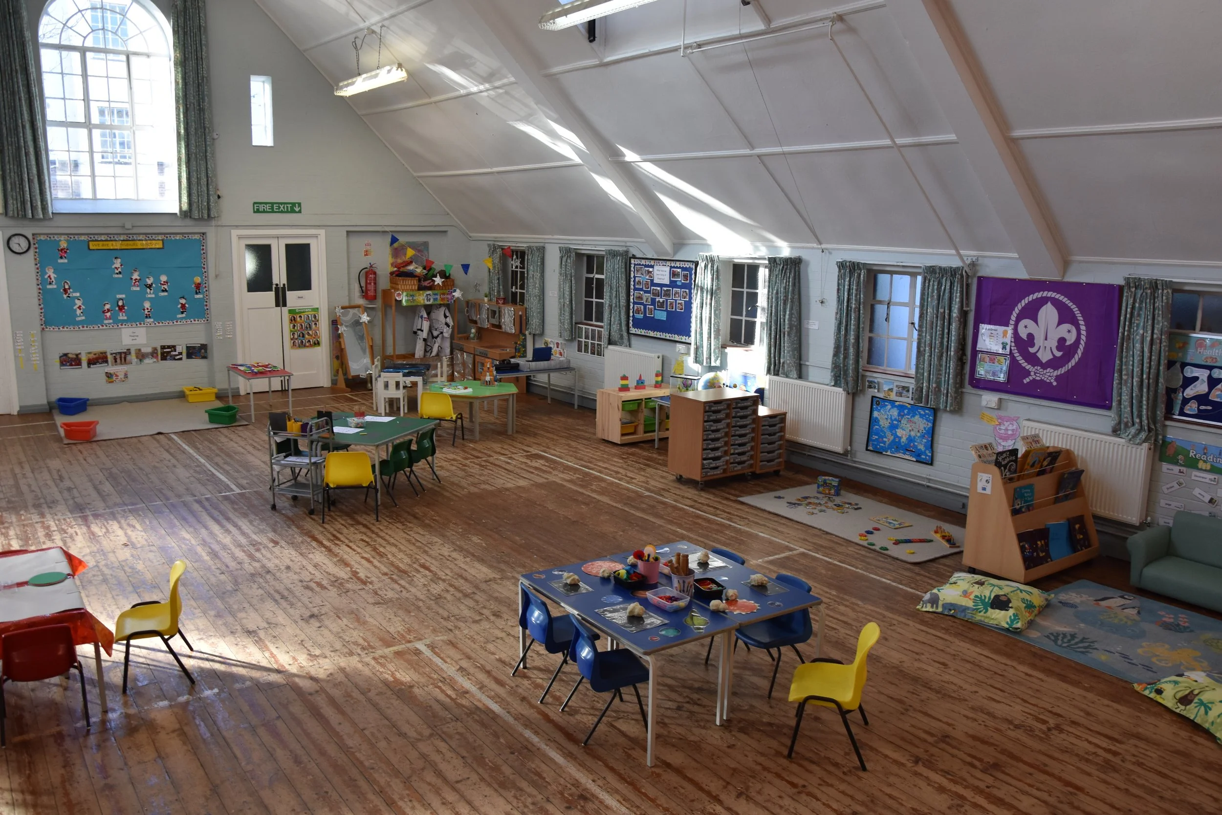 A large, empty classroom or playroom decorated with colorful toys, tables, and chairs, featuring a wooden floor, windows with curtains, and various bulletin boards and posters on the walls.