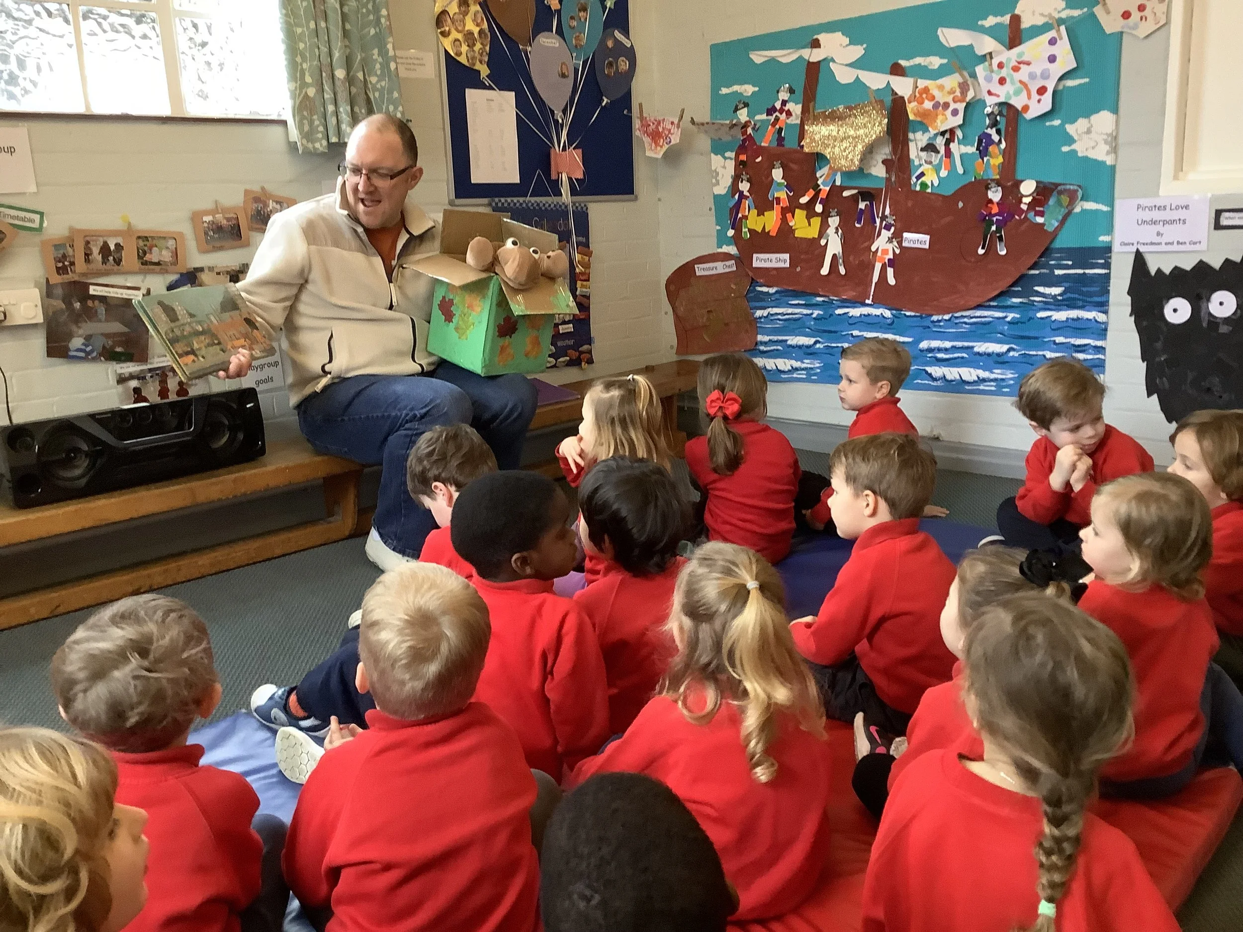 A man sitting on a bench reading a storybook to a group of children dressed in red uniforms, with a colorful pirate-themed mural in the background.