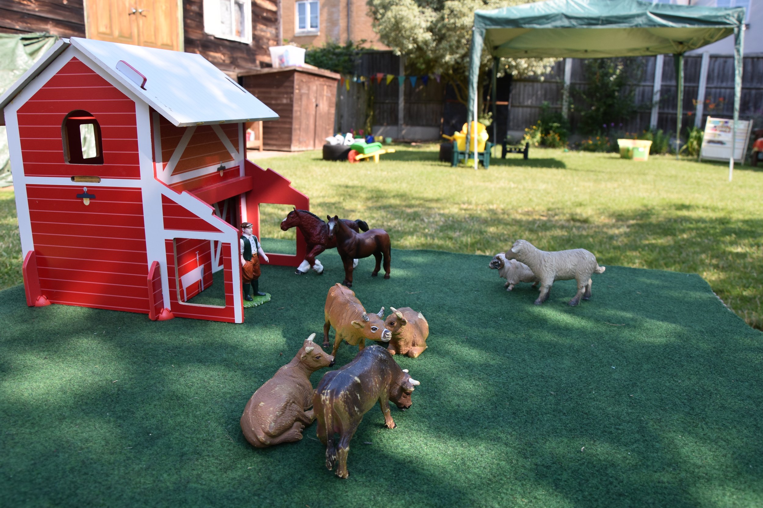 Children's toy farm scene with a red barn, plastic animals including horses, cows, sheep, and a person figure, set on a green surface in a backyard with grass, trees, and outdoor furniture.