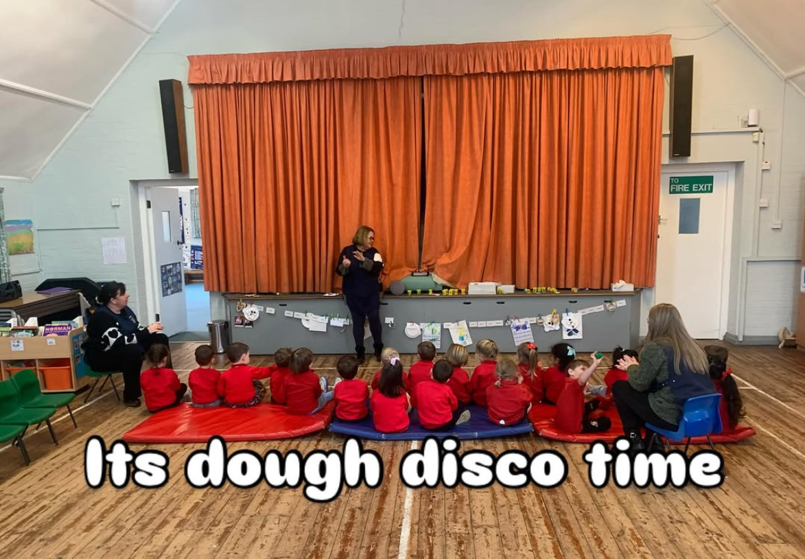 A group of children in red shirts sitting on mats watching a puppet show on a stage with an orange curtain. Two adults are present, one performing and the other sitting near the children. The caption says, "Its dough disco time."