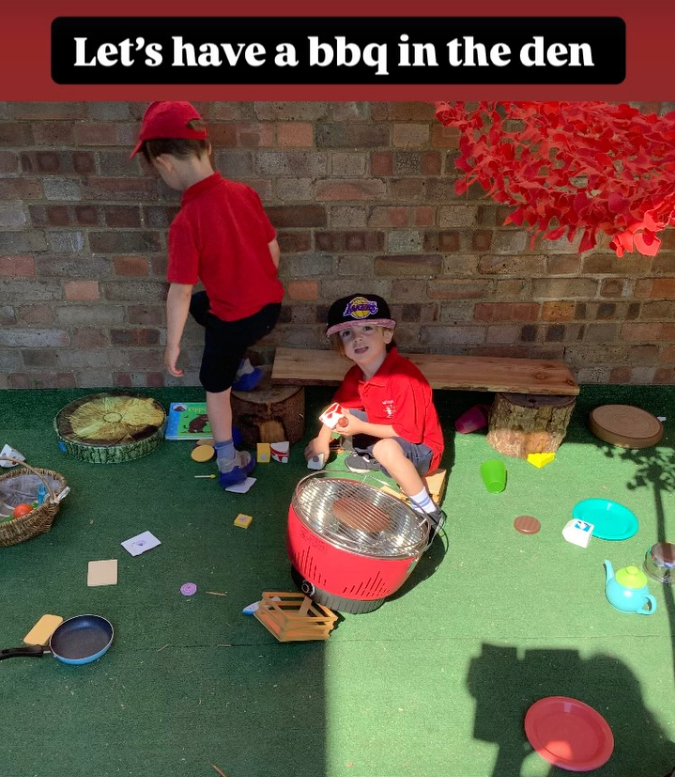 Two young boys playing with toy barbecue and outdoor play kitchen items in a backyard. One boy is sitting on a stool, holding a toy burger, while the other stands nearby, wearing a red cap. There are various toys scattered on the artificial grass, a brick wall in the background, and a sign at the top that reads 'Let's have a BBQ in the den.'