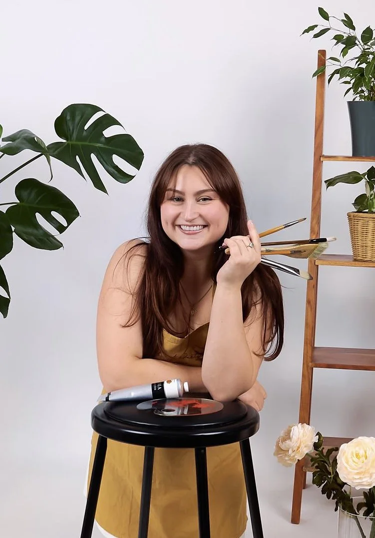 A woman with long brown hair smiling, holding paintbrushes, sits behind a black turntable with a vinyl record, surrounded by houseplants and flowers.