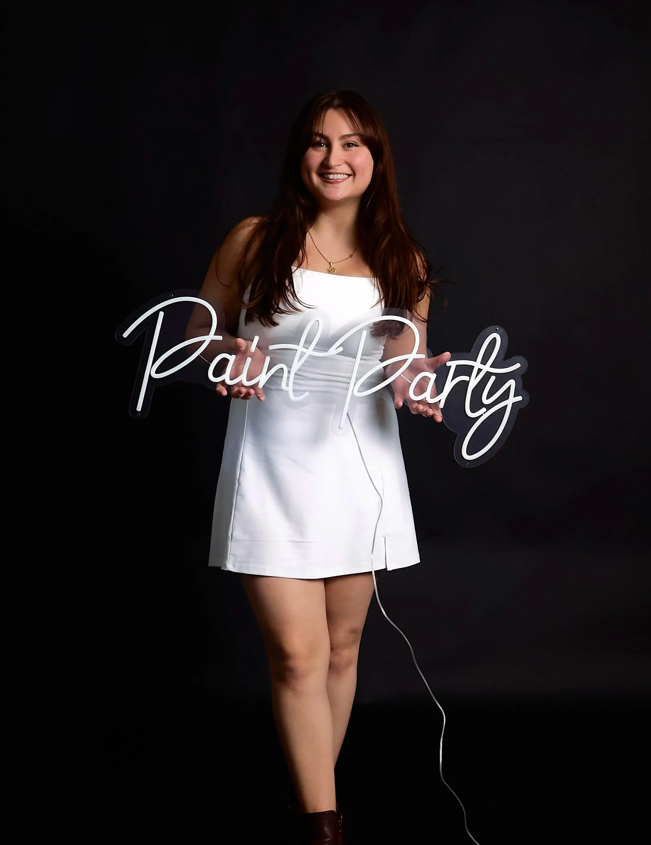A woman smiling in a white dress holding a sign that reads 'Paint Party' against a black background.