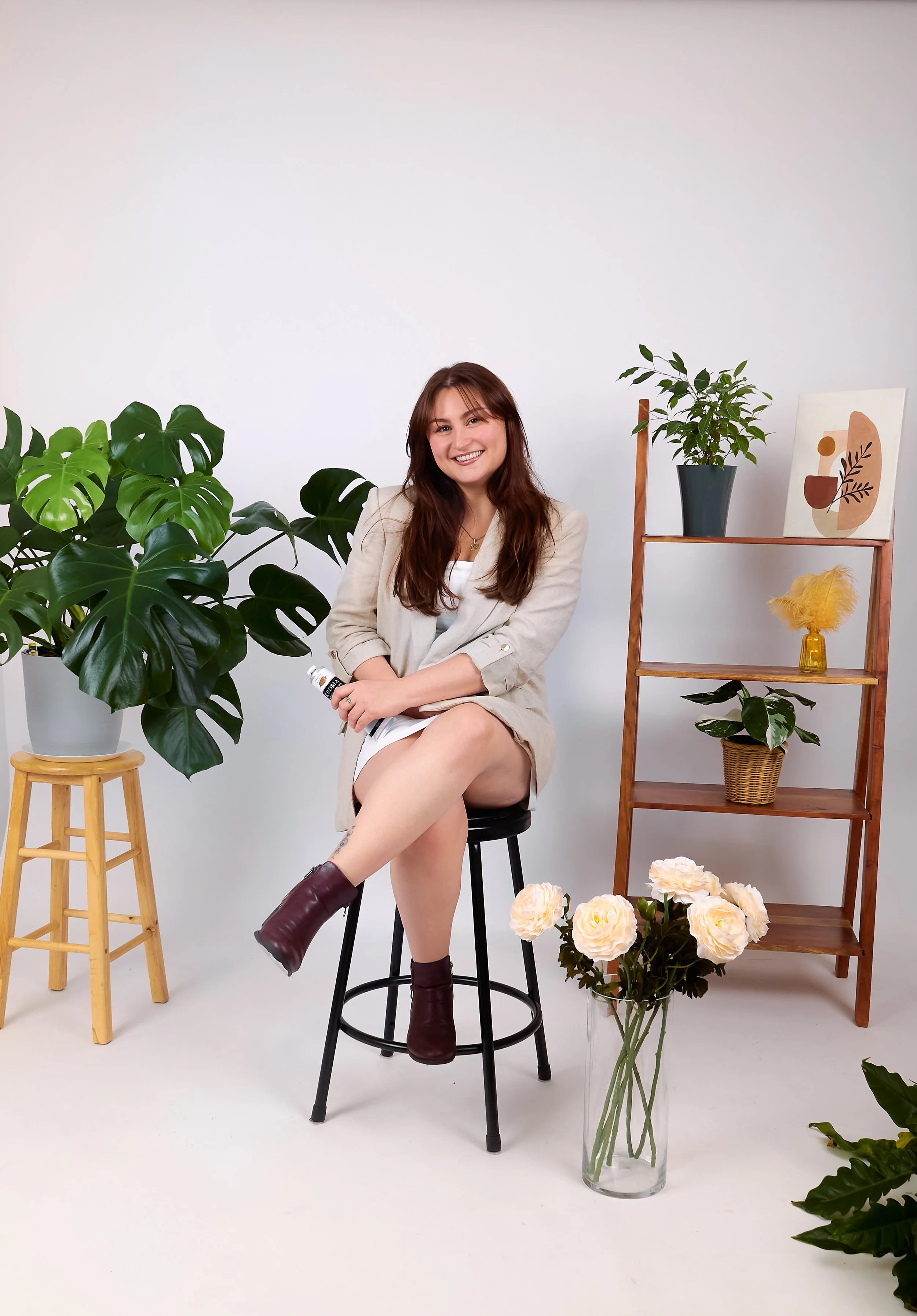 A young woman with long brown hair and a beige blazer sitting on a black stool, smiling, surrounded by houseplants and flowers in a white studio. She is holding a small white bottle in her left hand, her right ankle is resting on her left knee. The background and floor are white, with a wooden shelf to her right containing a potted plant, a framed abstract art piece, and a yellow feathered decorative item. A vase of white flowers is on the floor in front of her.