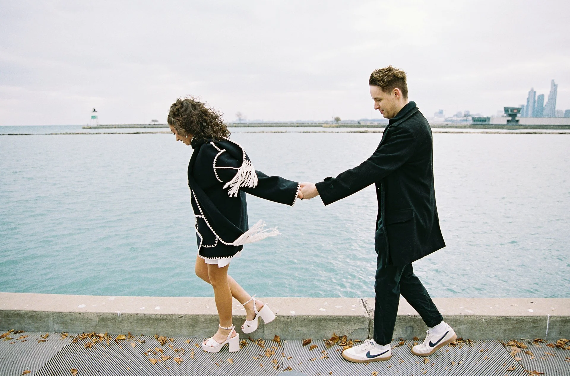 A man is pulling a woman by her arm along a waterfront promenade with water and city skyline in the background.