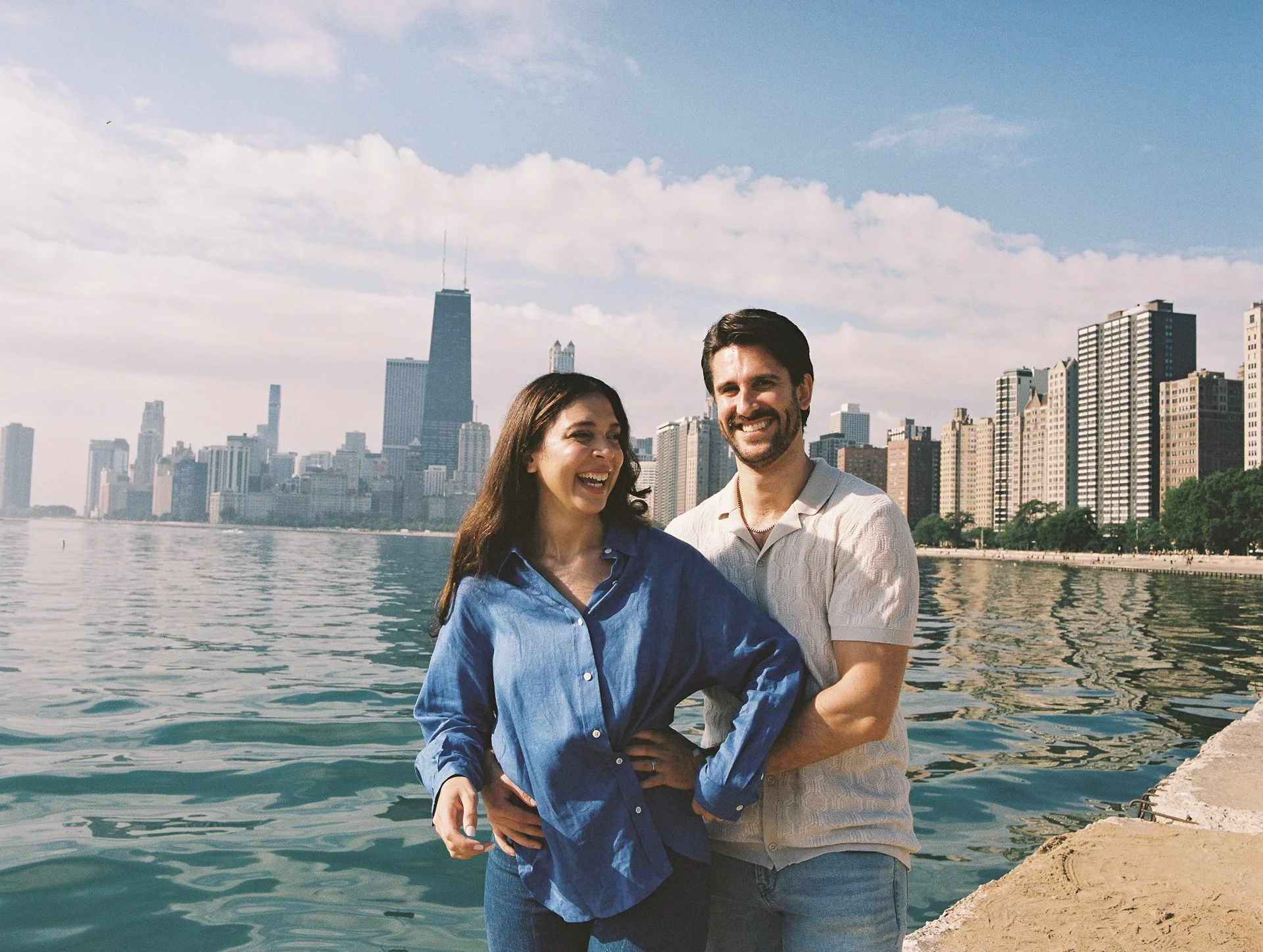 A smiling couple standing by the waterfront with the Chicago skyline in the background, holding each other and enjoying a sunny day.