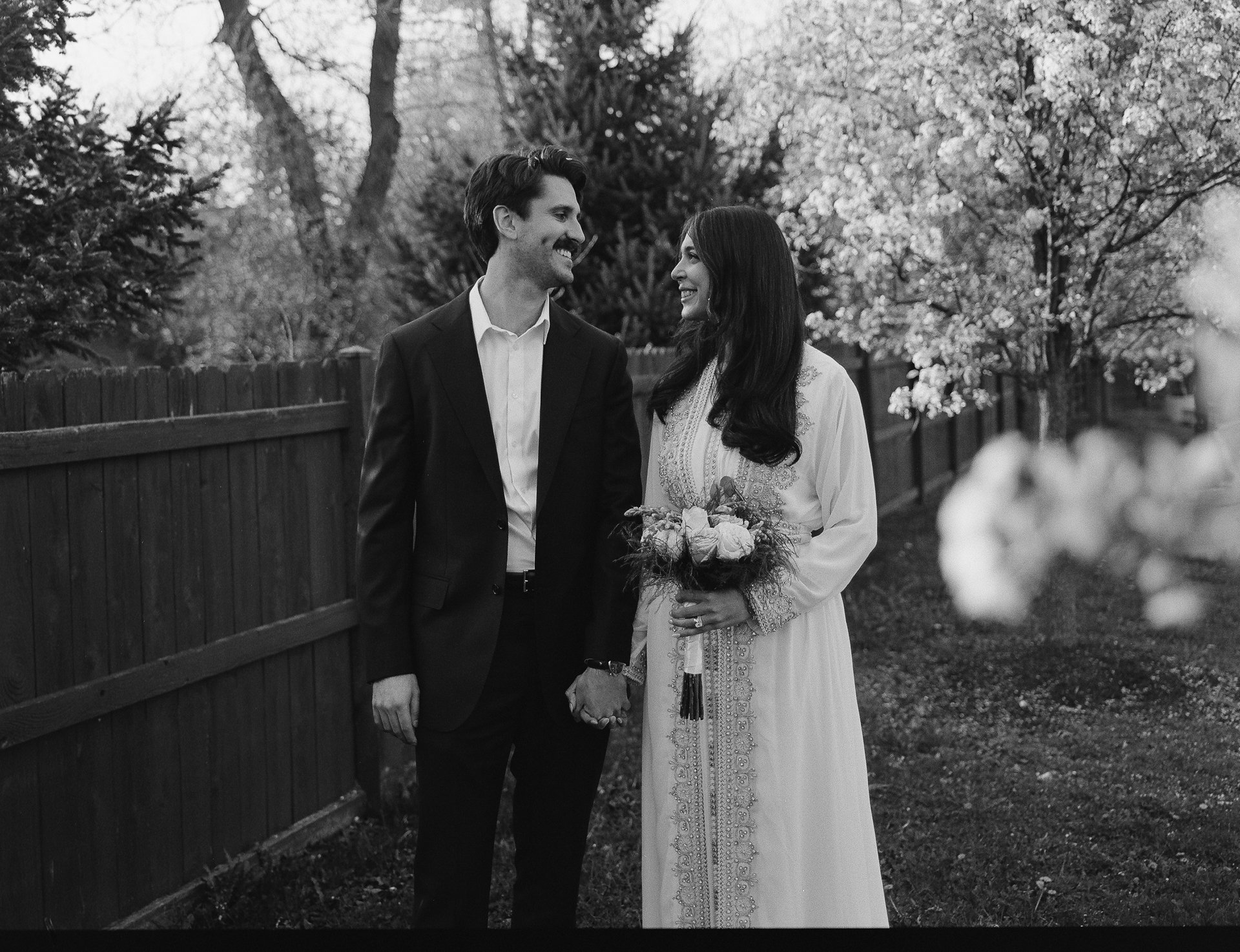 A black and white photo of a couple holding hands outdoors, smiling at each other. The man wears a dark suit and white shirt, and the woman wears a long white dress and holds a bouquet of flowers. They stand next to a wooden fence with trees in the b