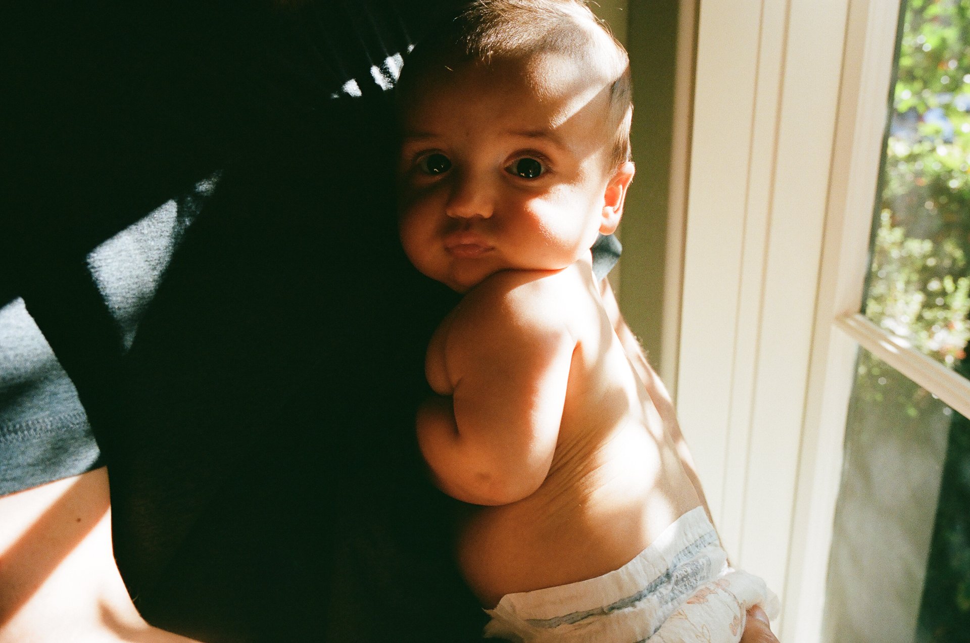 A young child with large, bright eyes, being held near a window with sunlight illuminating their face and body.