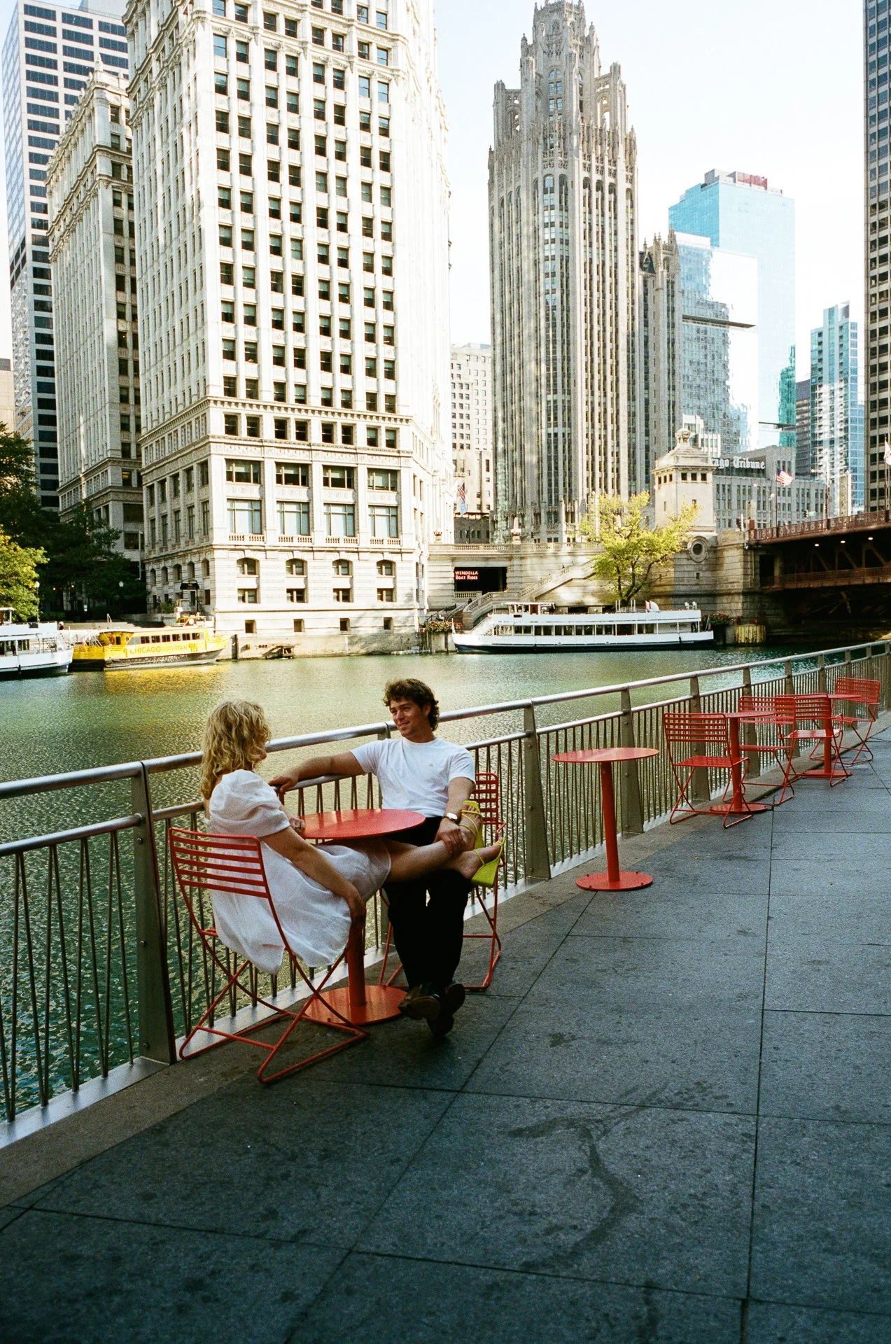 Two people sitting at a red outdoor table by the river in a city, with tall skyscrapers and boats in the background.