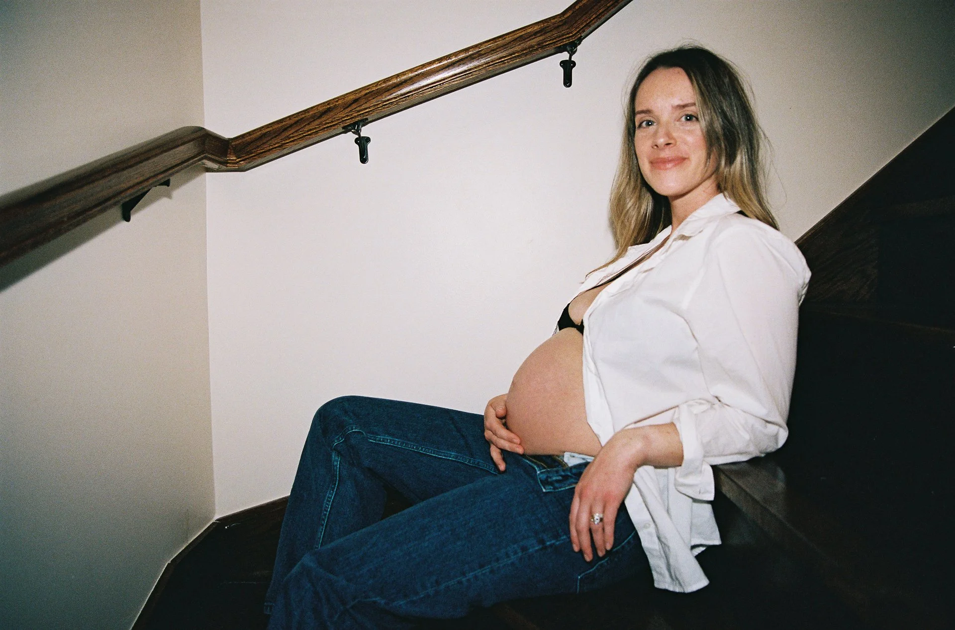 A pregnant woman sitting on a staircase, smiling at the camera, wearing a white shirt and jeans.