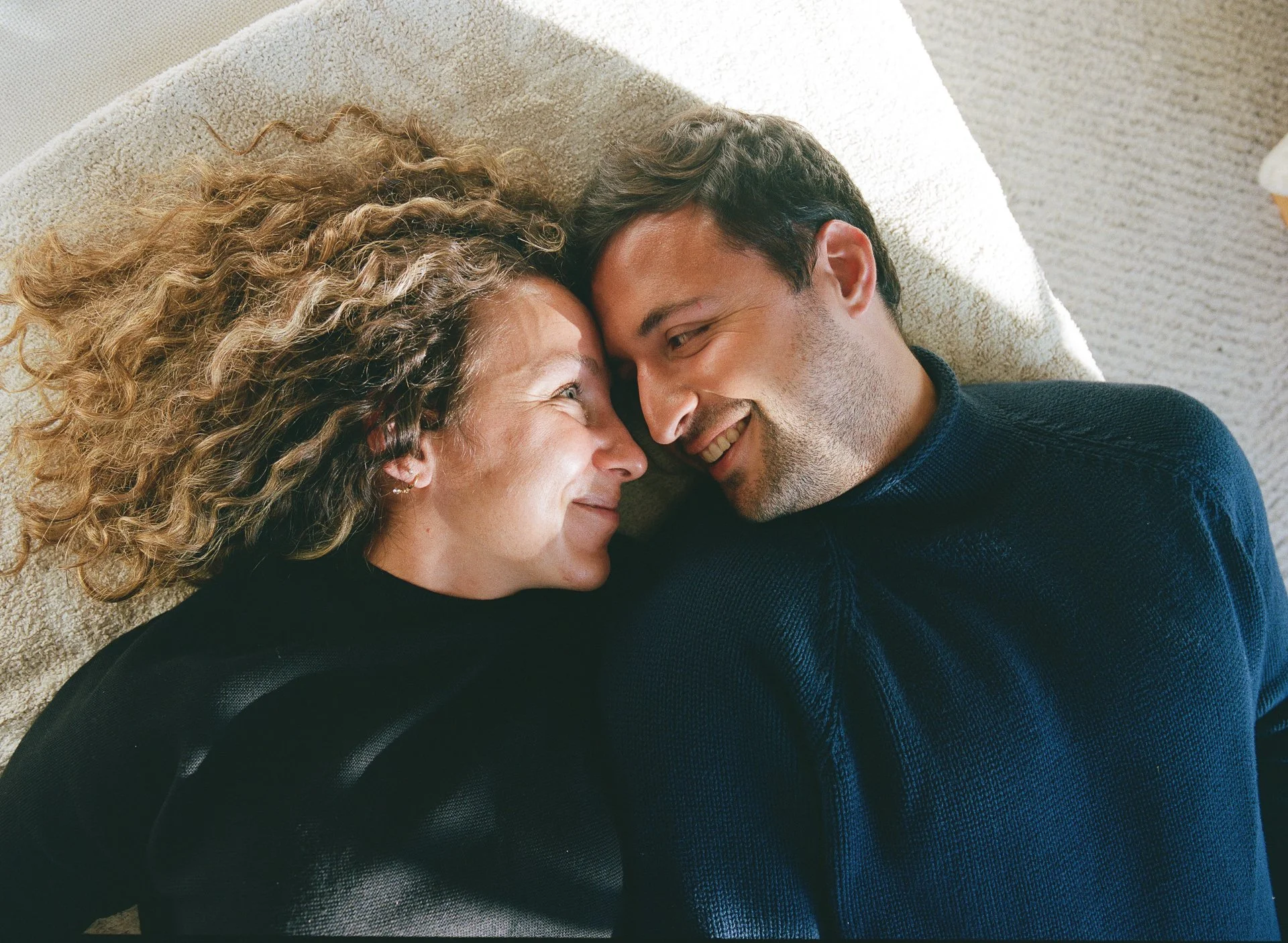 A woman and a man lying on a carpeted floor, forehead to forehead, smiling and looking at each other affectionately.