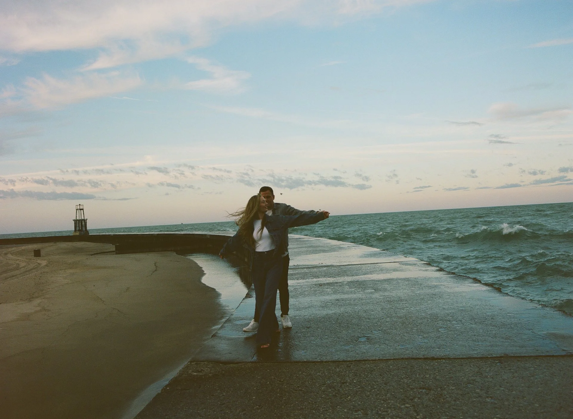 Two people, a man and a woman, standing on a wet concrete pier by the ocean, with a sky filled with clouds in the background.