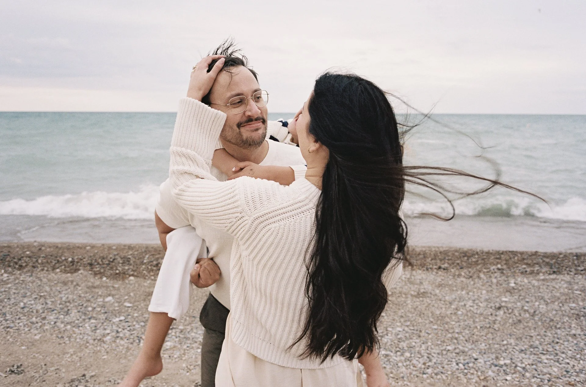 A couple embracing on a beach with the ocean in the background, the woman with long dark hair and the man with glasses and facial hair, both wearing light-colored sweaters.