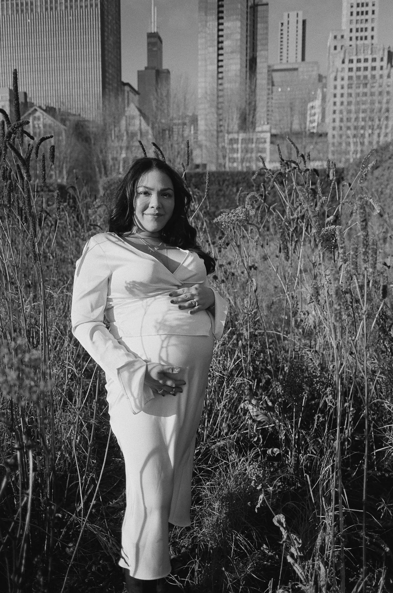 A pregnant woman standing in a field of tall grass and wildflowers with a city skyline in the background, in black and white.