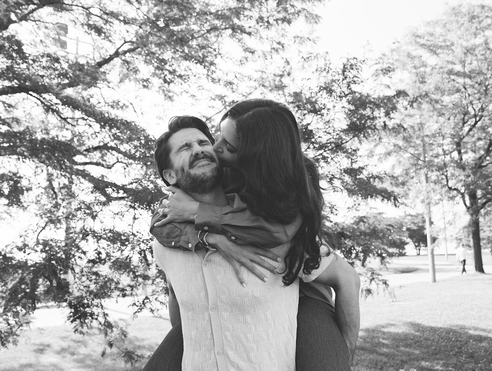 A woman with long dark hair kisses a man with dark hair on the cheek while he smiles with eyes closed, during daytime in a park with trees in the background.