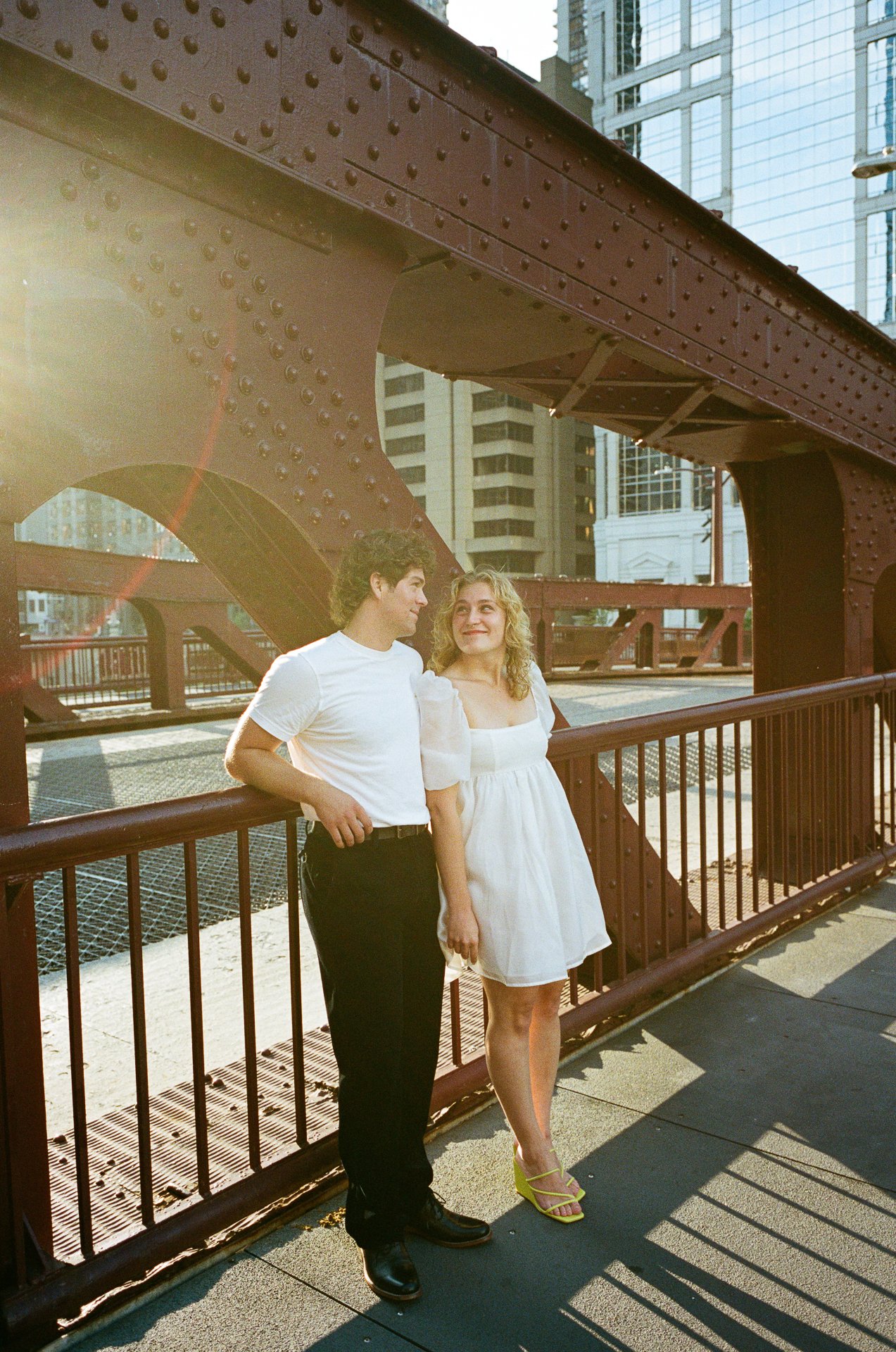 A young man and woman standing together on a city sidewalk under a bridge, with tall buildings in the background, sunlight shining on them, and the woman wearing a white dress and yellow high heels.
