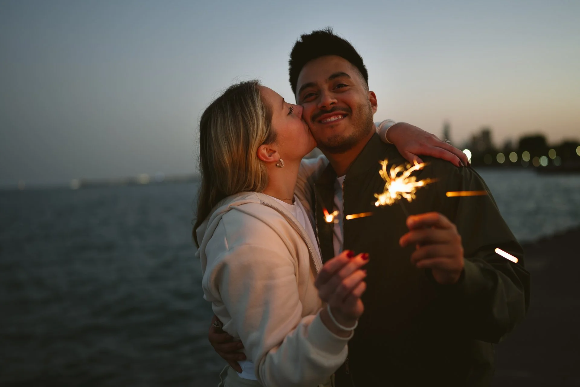 A couple embracing by the water at dusk, with one holding a lit sparkler and the woman giving a kiss on the cheek to the man.