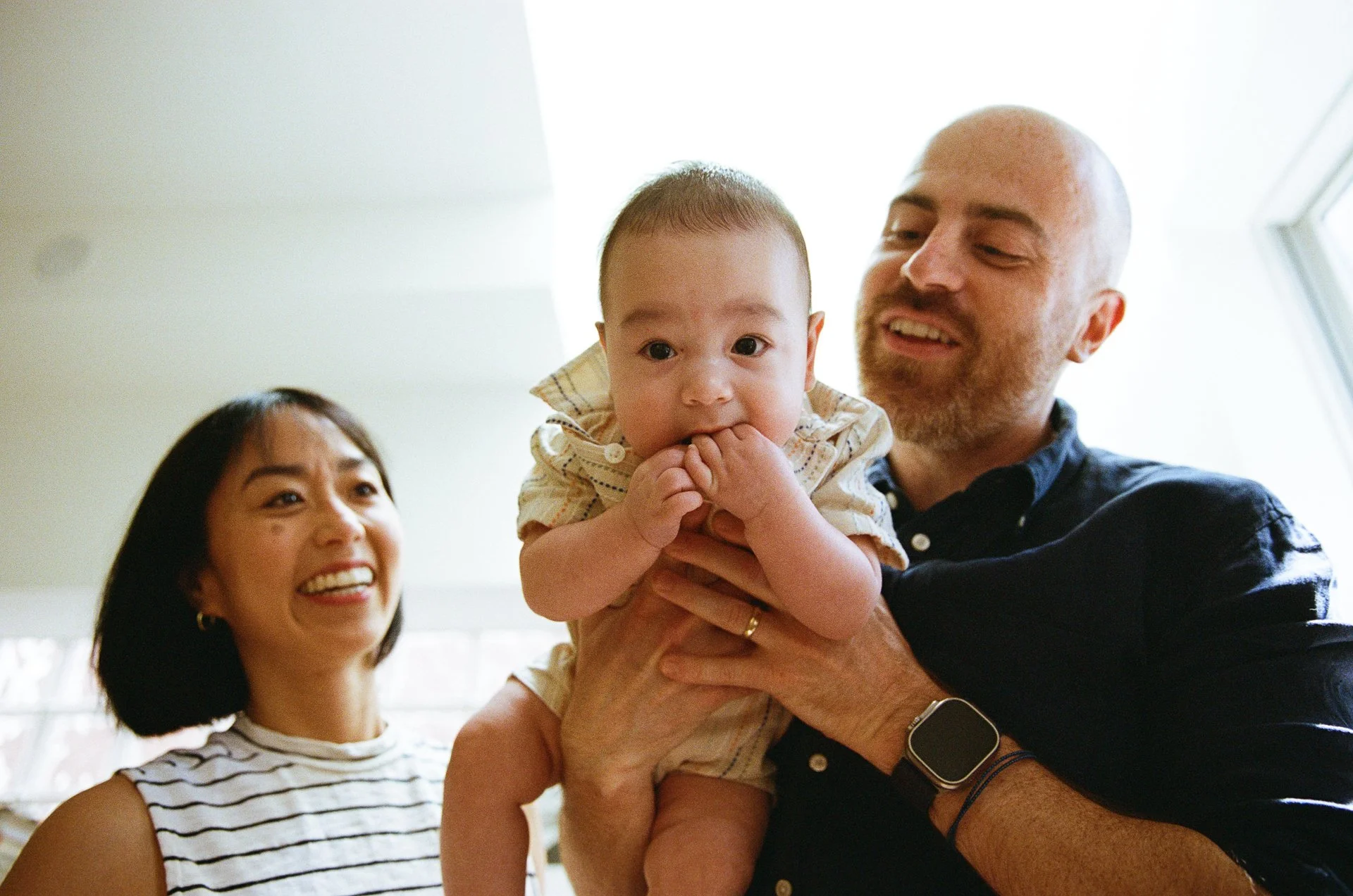 A smiling woman, a man with a beard holding a baby, in a well-lit room with a window.