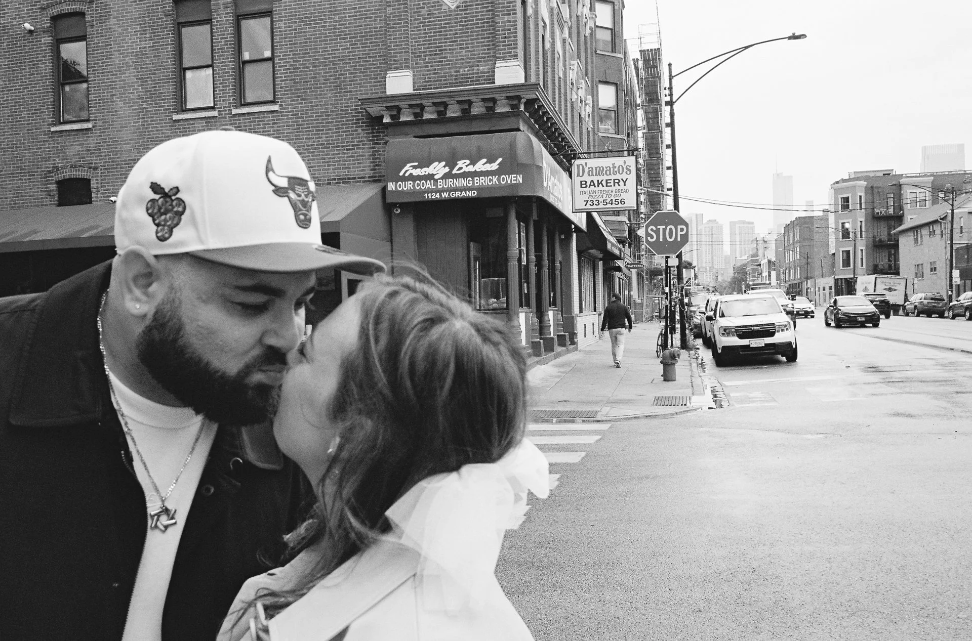 A couple sharing a kiss on a city sidewalk, with storefronts, cars, and a street scene in the background.