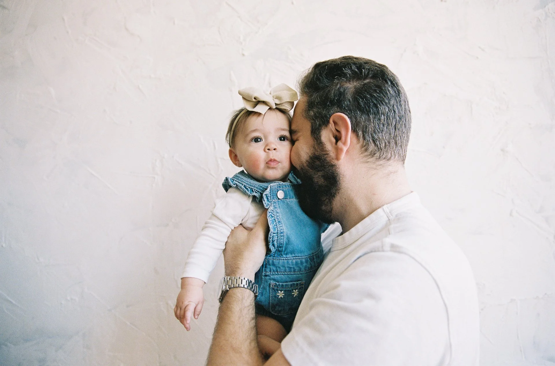 A man with a beard holding a young girl close, appearing to kiss her on the cheek against a plain white wall