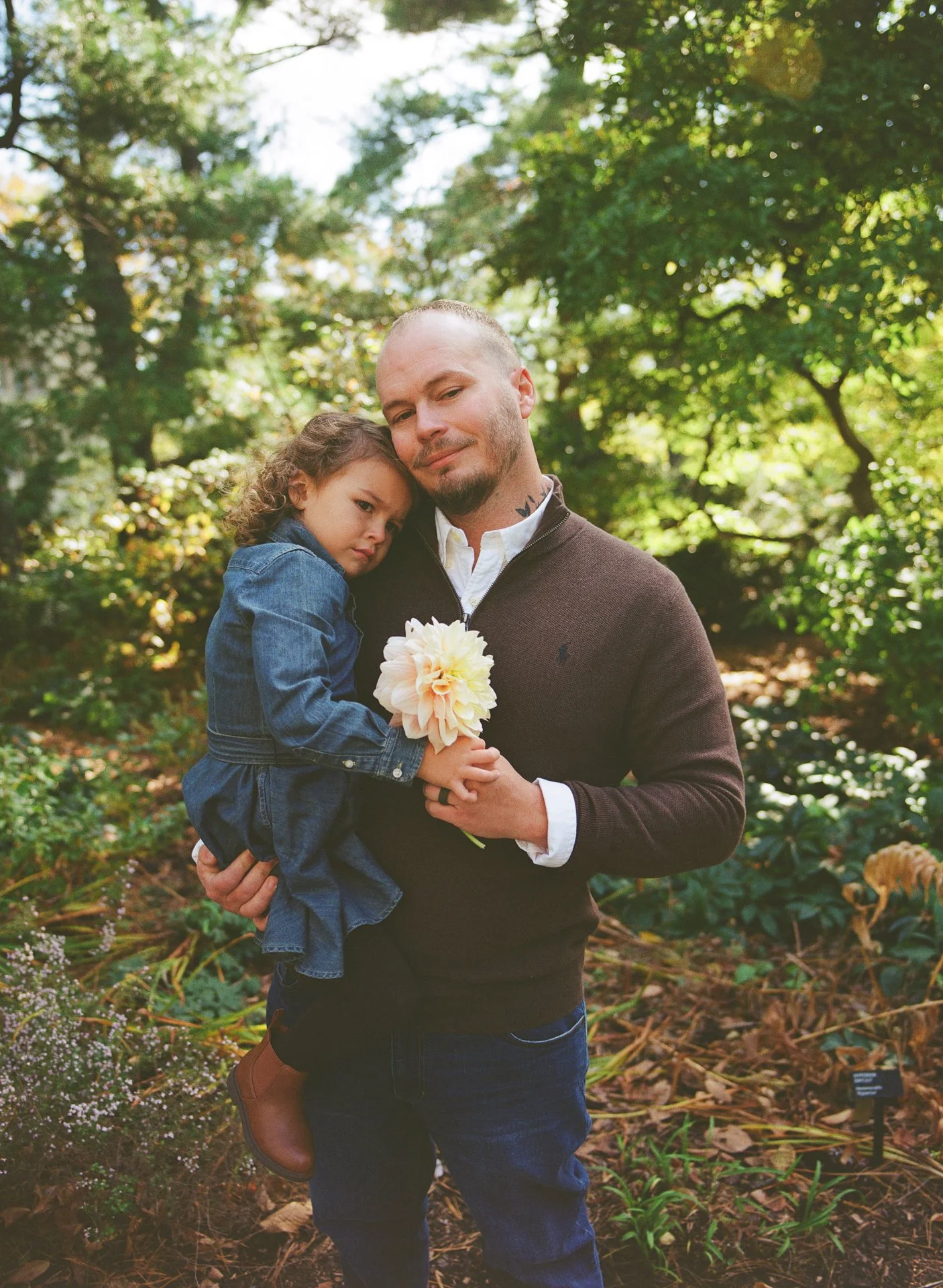 A man holding a young girl with curly hair and a denim dress in a forest setting, with trees and greenery in the background, and the girl holding a flower.
