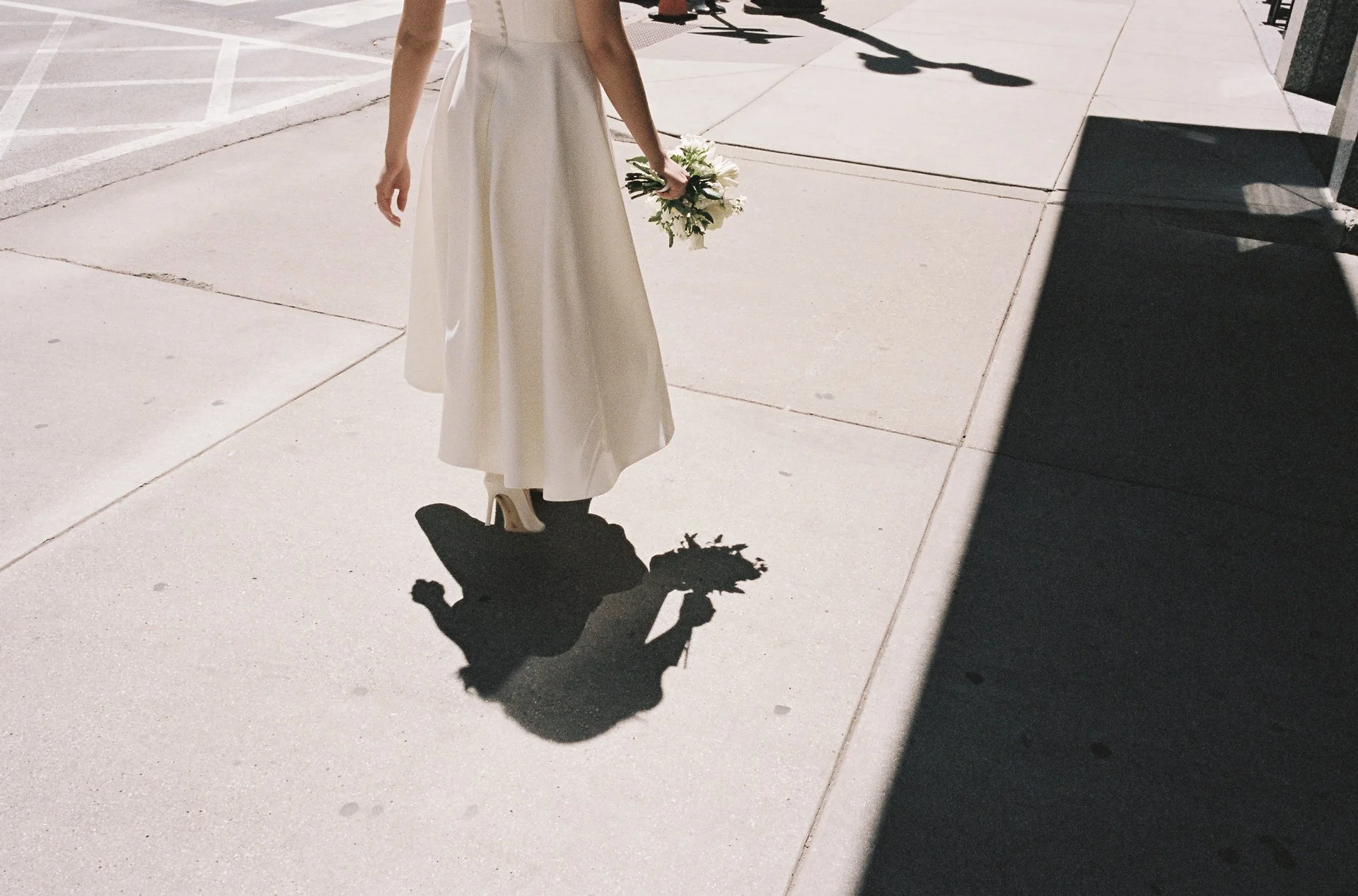 A woman standing on a sidewalk holding a bouquet of white flowers, with her shadow cast on the ground, in bright sunlight.