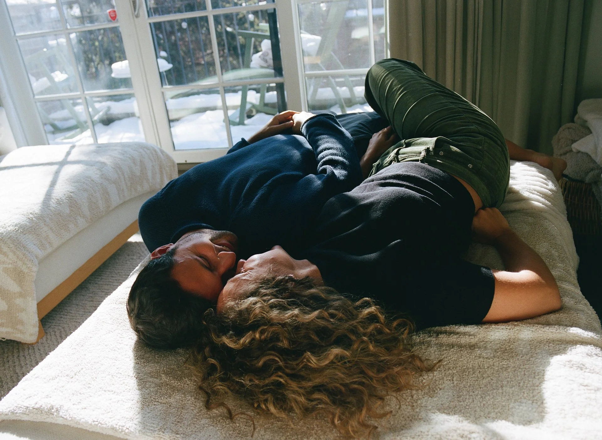 A man and woman lying on a bed, kissing, with sunlight coming through a glass door behind them.