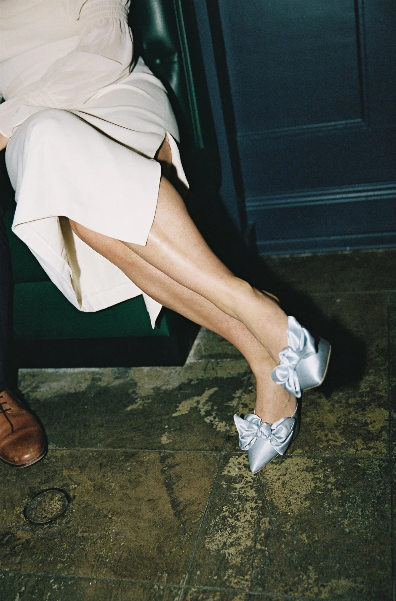 A person's legs and feet with silver satin shoes with large bows, sitting on a green bench. The person is wearing a cream-colored dress with black piping, and the background shows a dark wall and tiled floor.