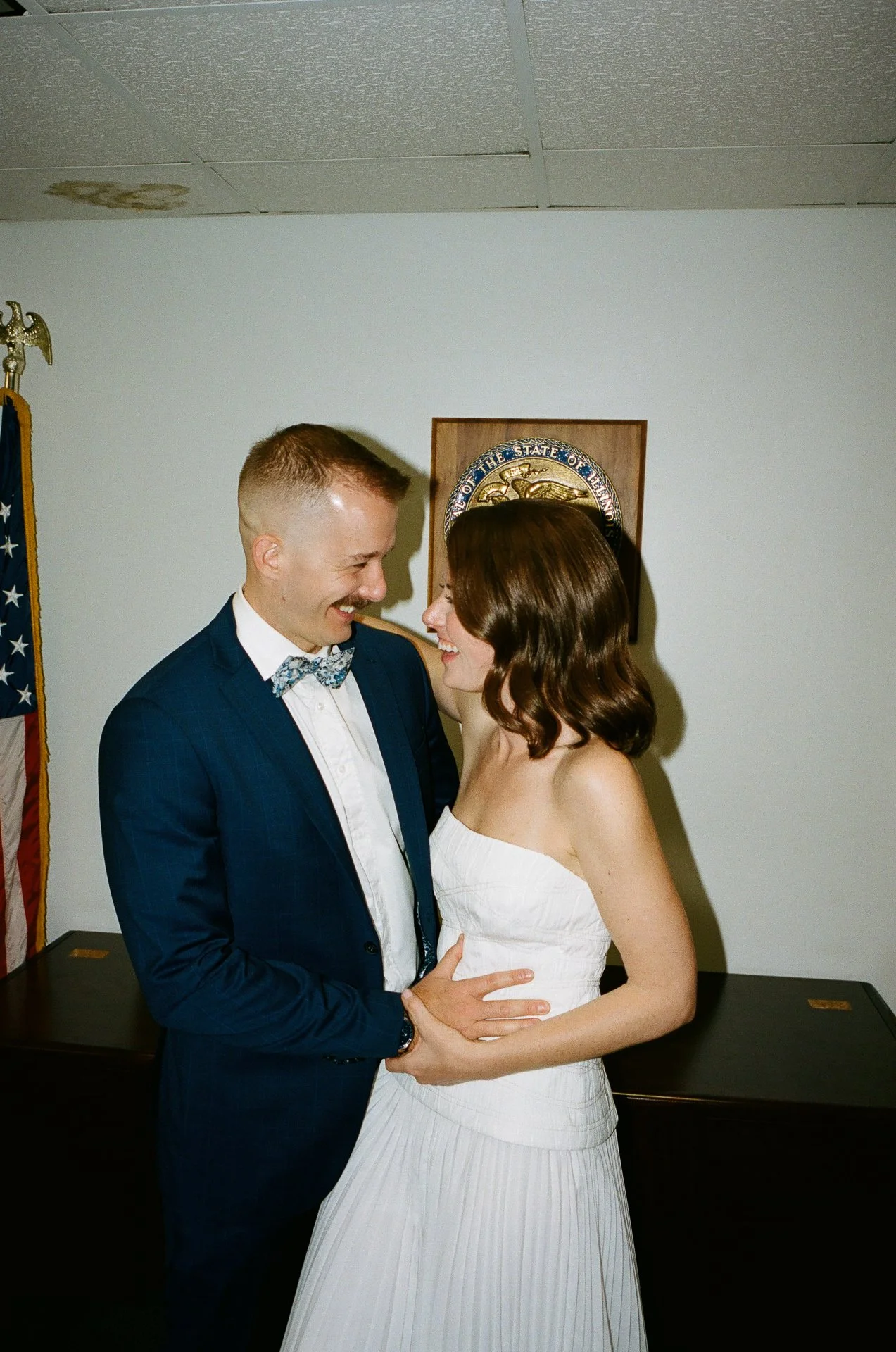 A couple dancing and smiling at their wedding, with the man wearing a tuxedo and the woman in a strapless white wedding dress, in a room with an American flag and a government seal on the wall.