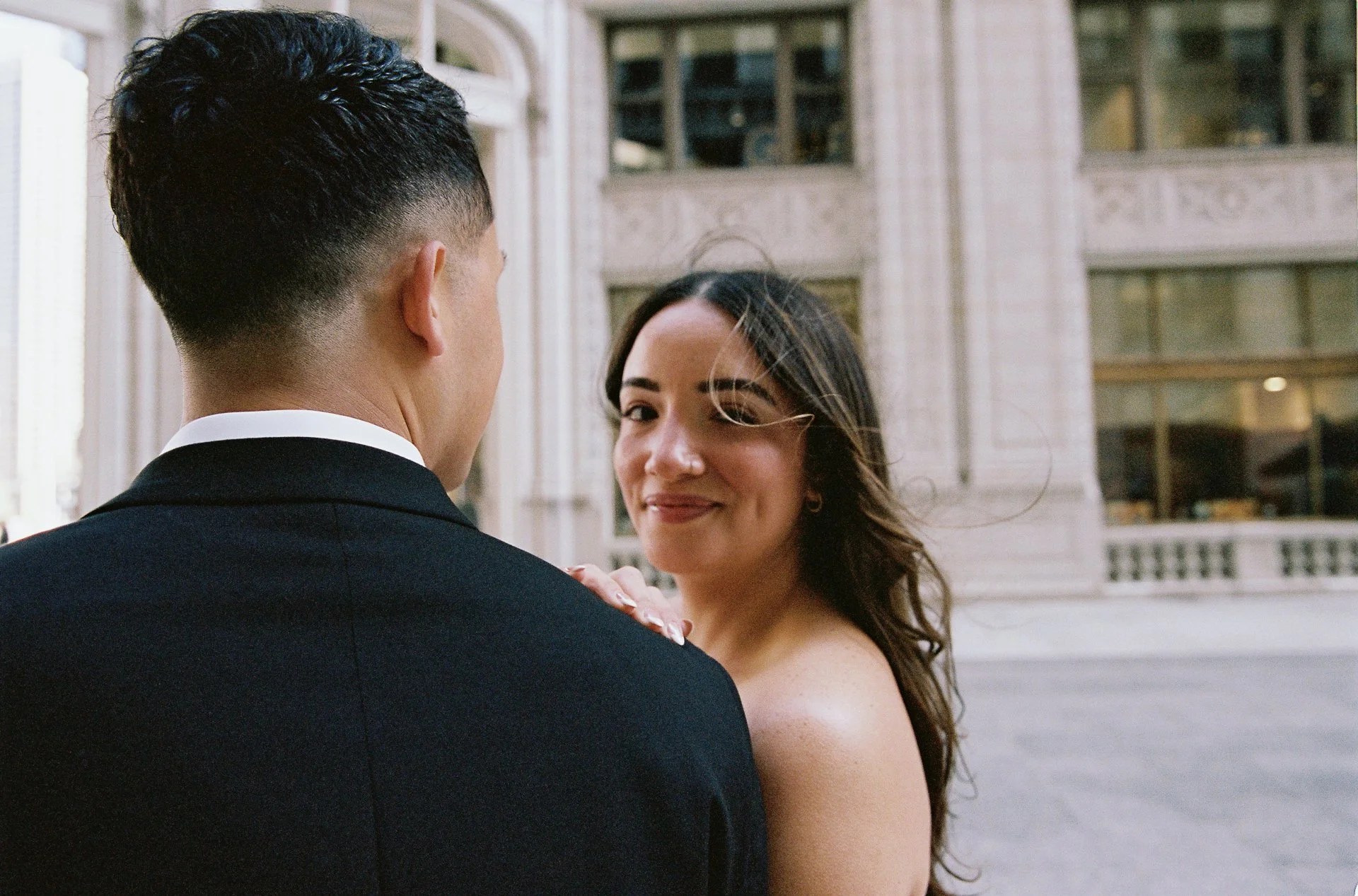 A couple dressed in wedding attire standing close together outdoors in front of a historic building, with the woman smiling and the man facing away from the camera.
